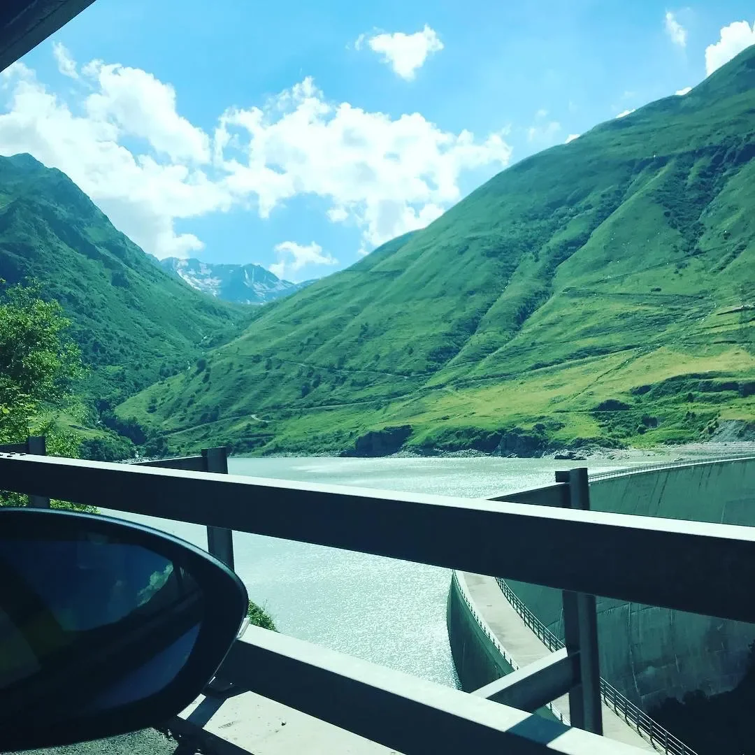 Scenic view of green mountains, a water reservoir, and a railing along a road, with a partly cloudy sky above.
