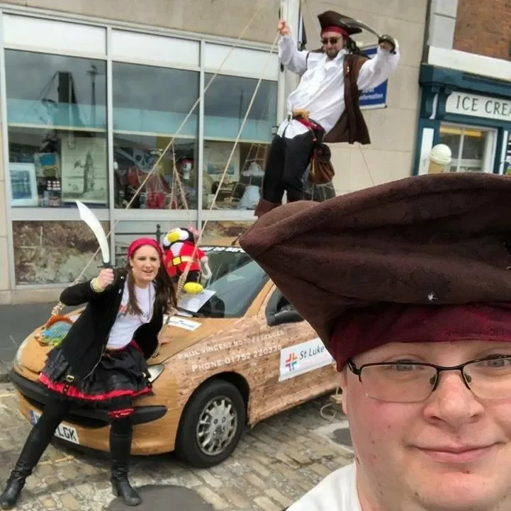 People dressed in Pirate costumes standing around a car, with one person sitting on the car and another standing on top, outside a building with ice cream shop.