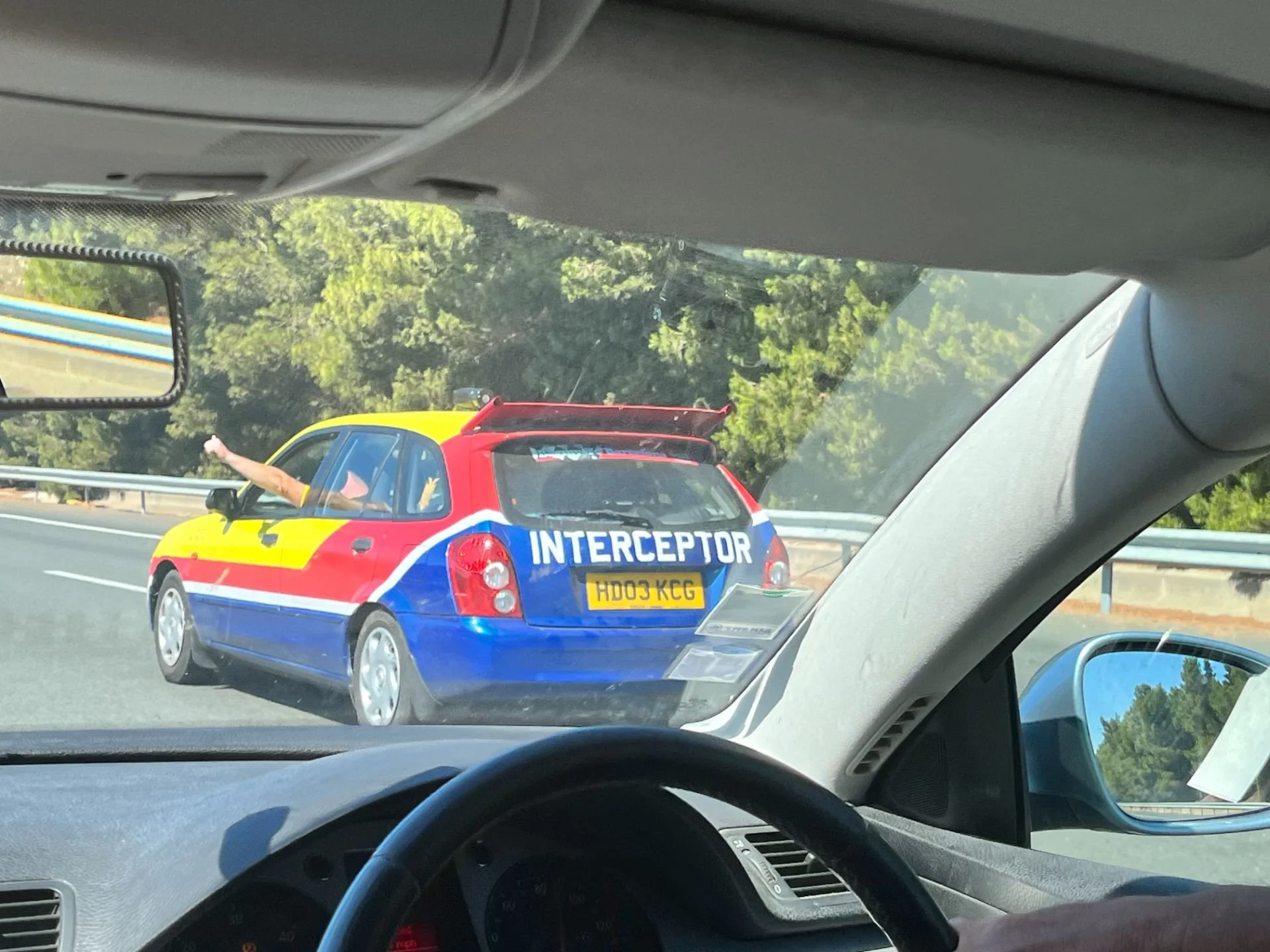 A toy car resembling an emergency responder vehicle with red, yellow, and blue colors, marked with the word 'INTERCEPTOR,' is seen on a highway through a car's windshield.