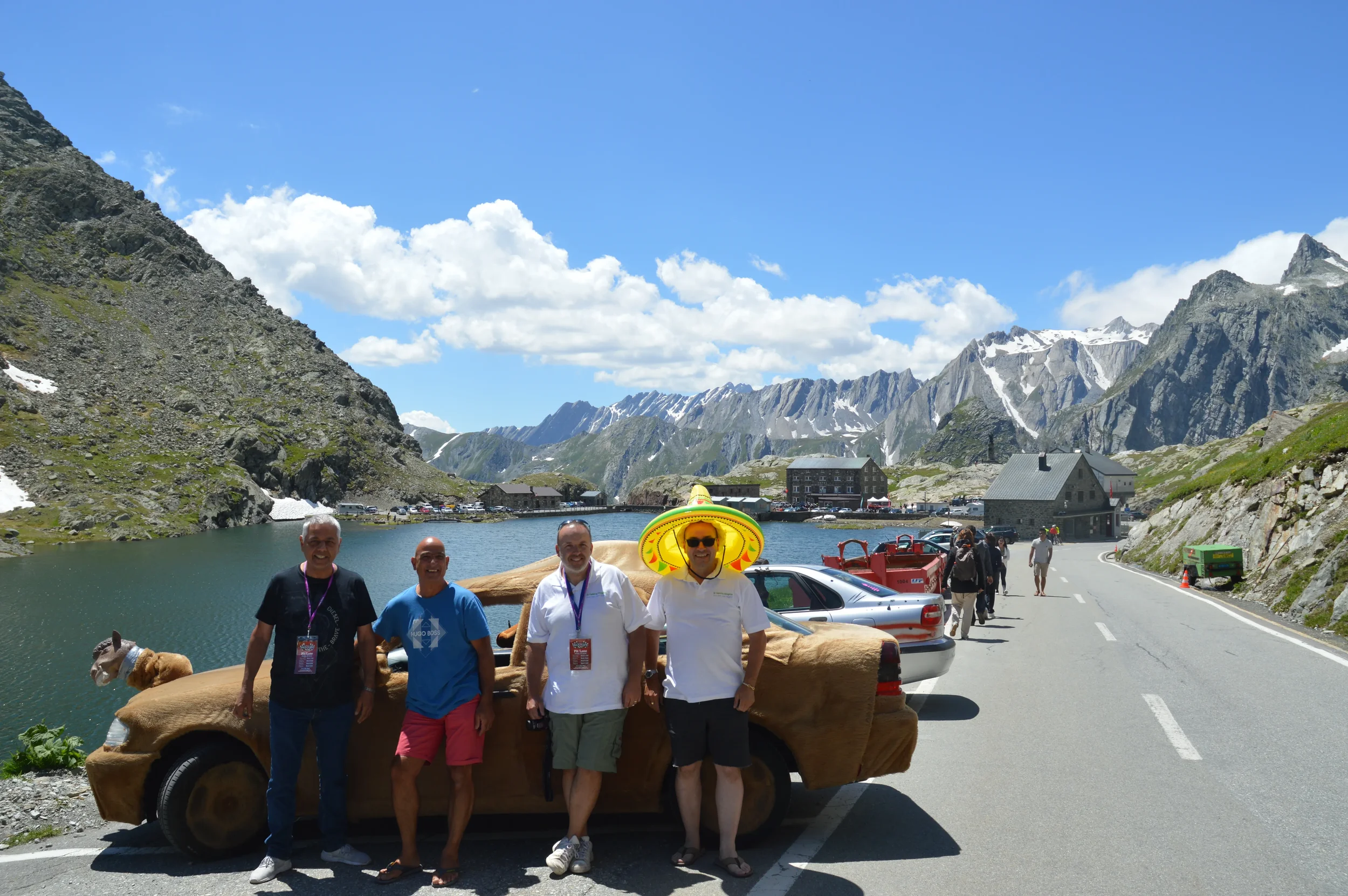 Four men standing in front of a car that is covered in brown material, with a lake, mountains, and a few buildings in the background. One man is wearing a large yellow sombrero and sunglasses. The scene is bright and sunny with a clear blue sky.