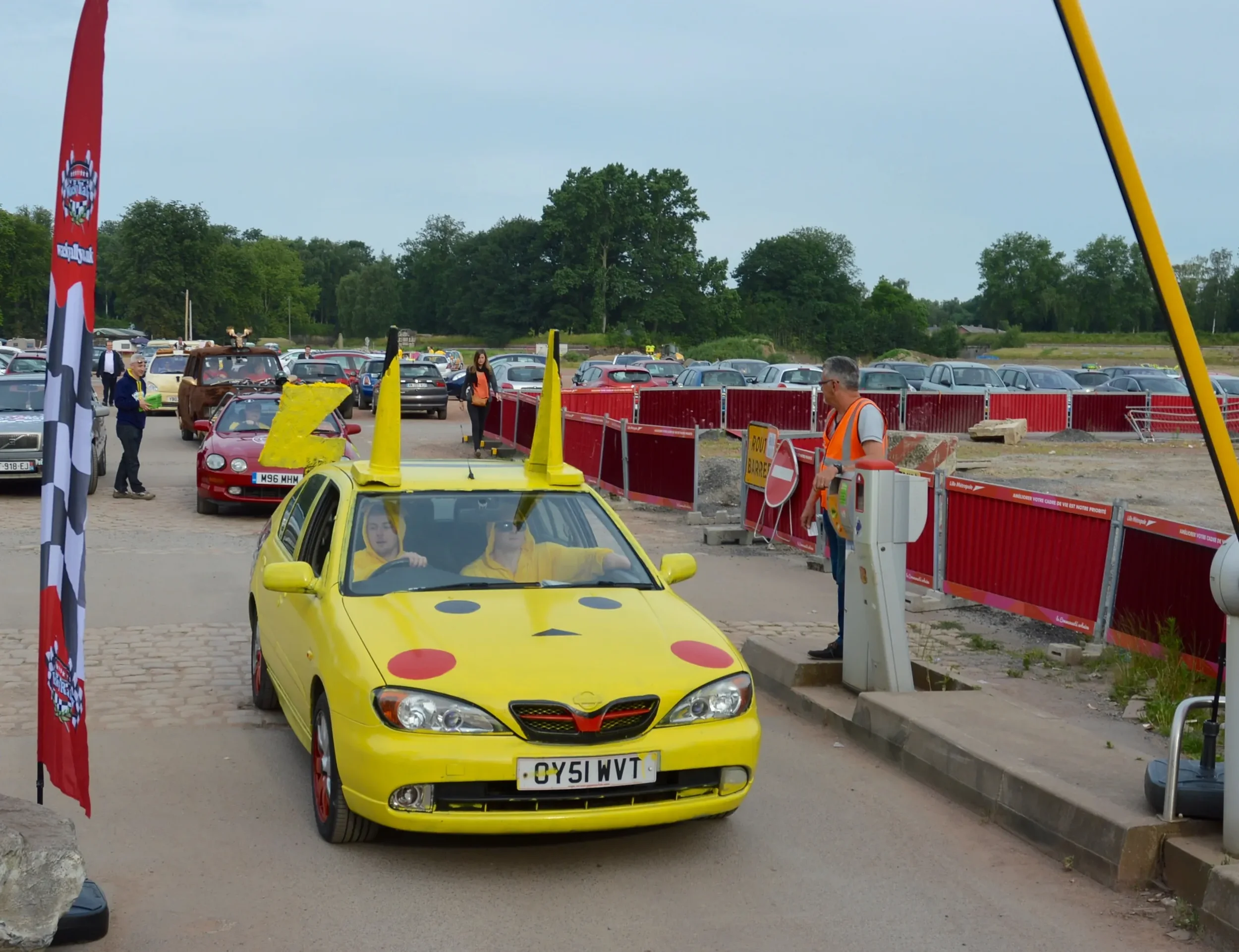 A yellow car decorated to look like Pikachu from Pokémon, with Pikachu face and ears on the roof, entering a parking lot with other cars and people around.