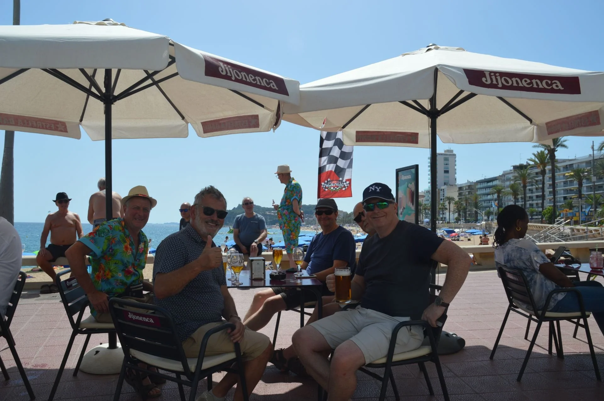 Group of people sitting at an outdoor beachside restaurant under umbrellas, enjoying drinks on a sunny day with a beach, ocean, and buildings in the background.