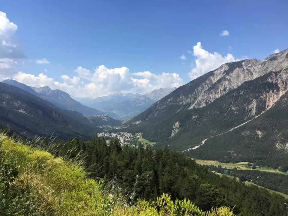 Scenic view of a mountain valley with green forests, rocky mountains, and a small town or village in the distance under a blue sky with scattered clouds.