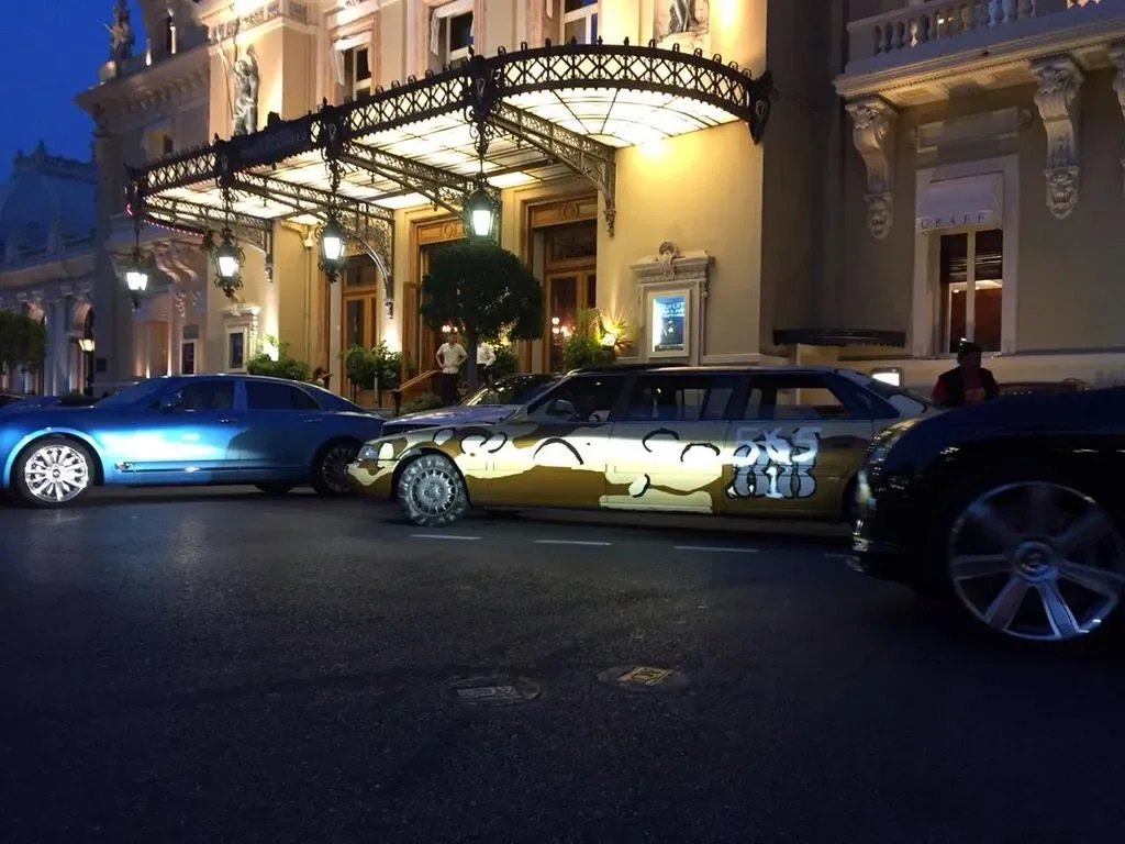 Luxury hotel with elaborate exterior lighting and ornate architecture, with several high-end cars parked in front, including a blue car, a gold car with graffiti, and a black car.