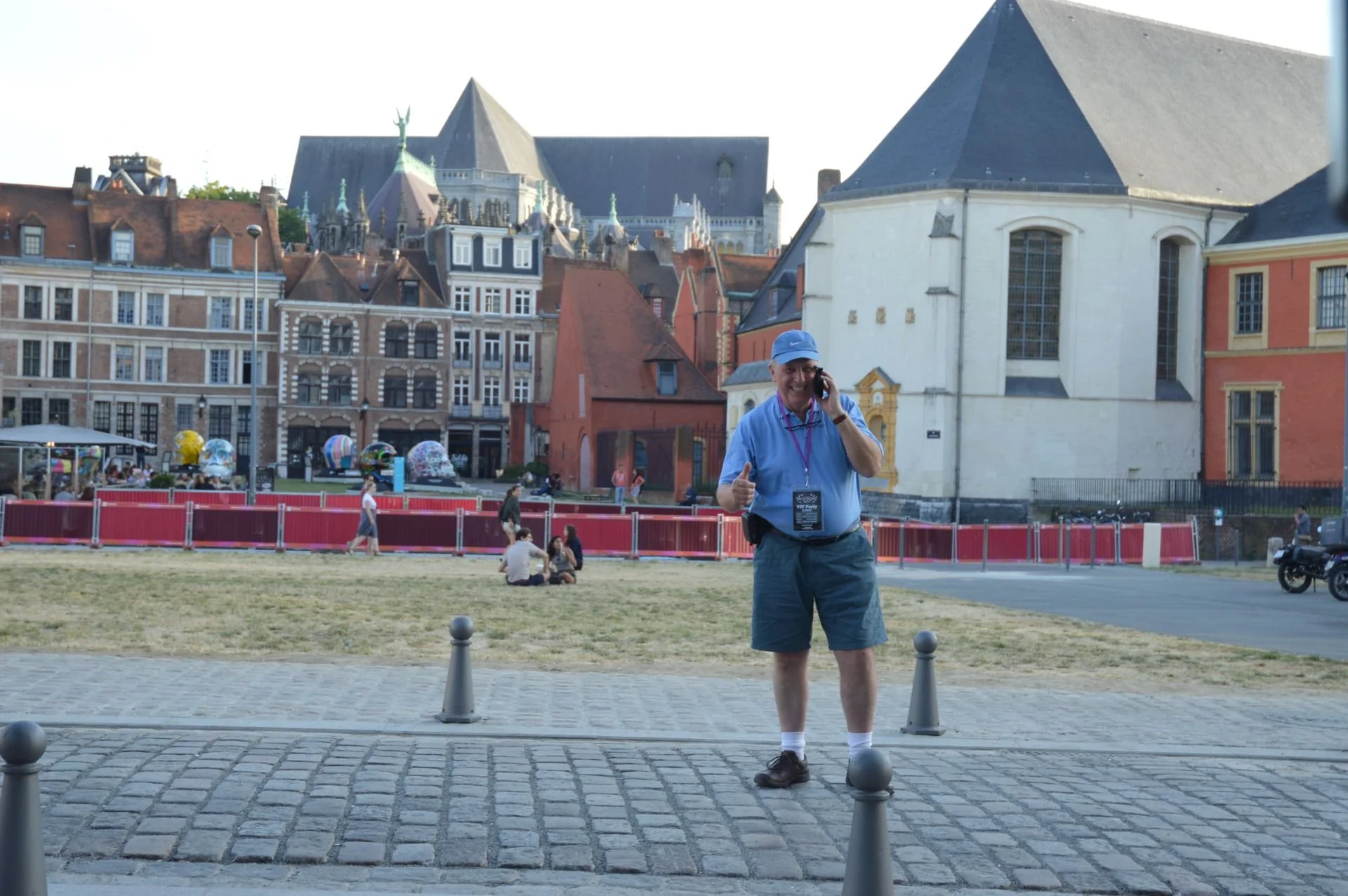 A man in casual clothing, including a blue cap and shorts, talking on a cellphone and giving a thumbs-up gesture, standing on a cobblestone street in a historic European city square with old buildings and a church in the background.