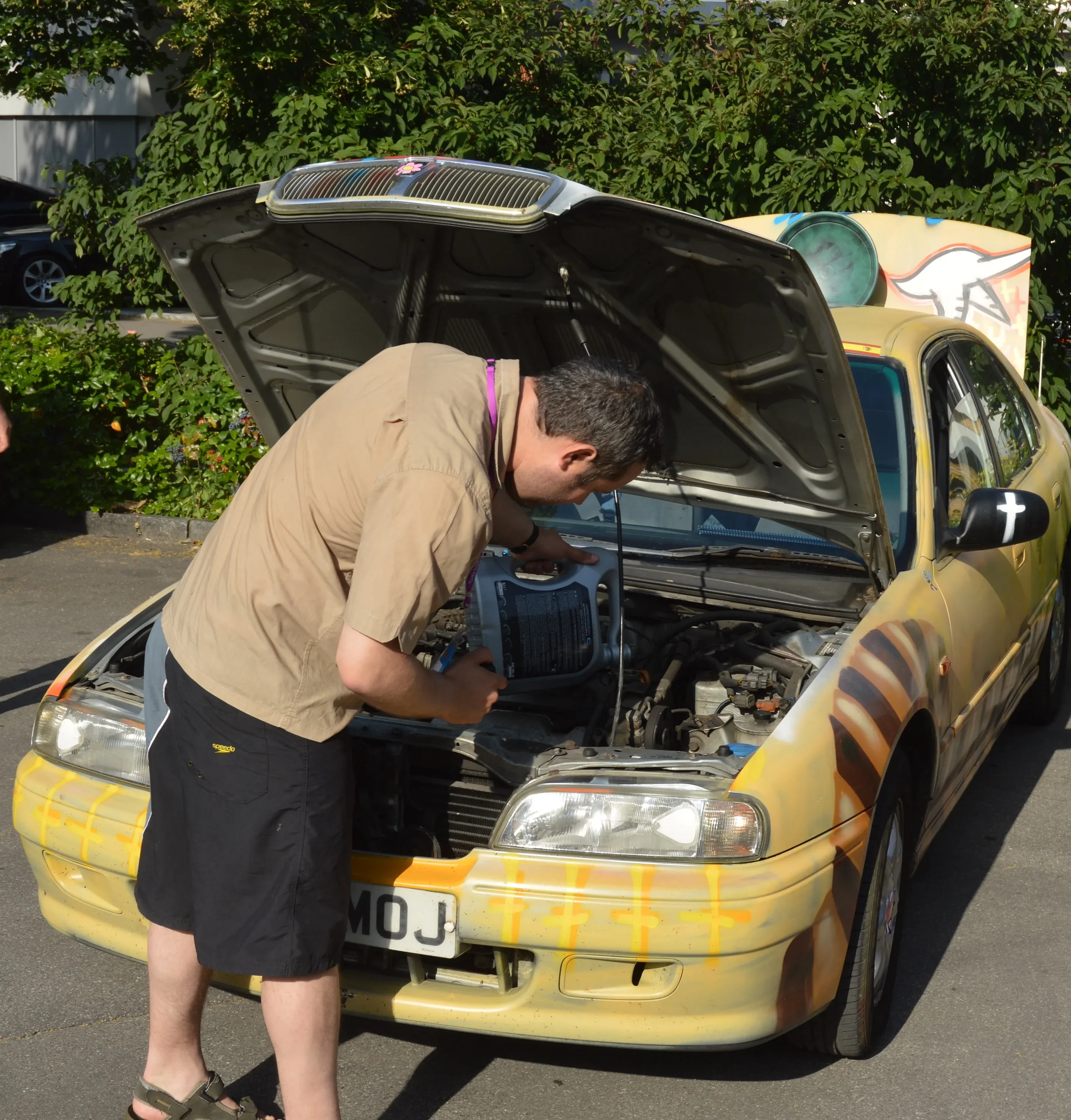 Man inspecting a yellow car with graffiti artwork on the hood, parked outdoors, with its hood open and engine exposed.