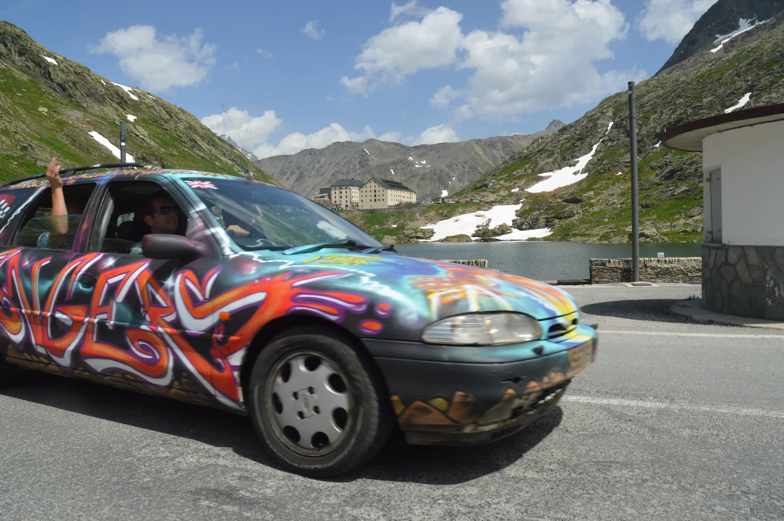 A colorful spray-painted car with a British flag sticker, driving on a mountain road near a lake under a partly cloudy sky.