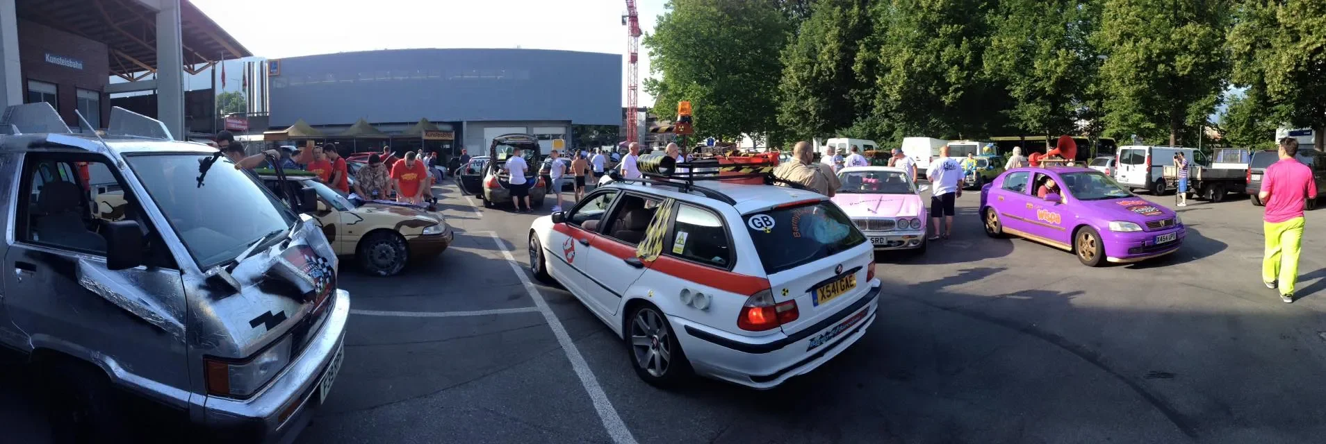 A parking lot filled with various small, colorful cars, some dressed up or decorated, and people walking around or standing near them, with trees and a large building in the background.