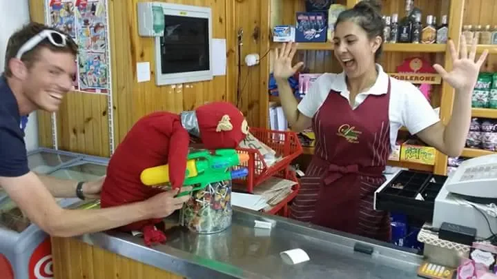 A man and a woman in a grocery store or convenience shop, jokingly playing with a stuffed red dog plush toy near the checkout counter.