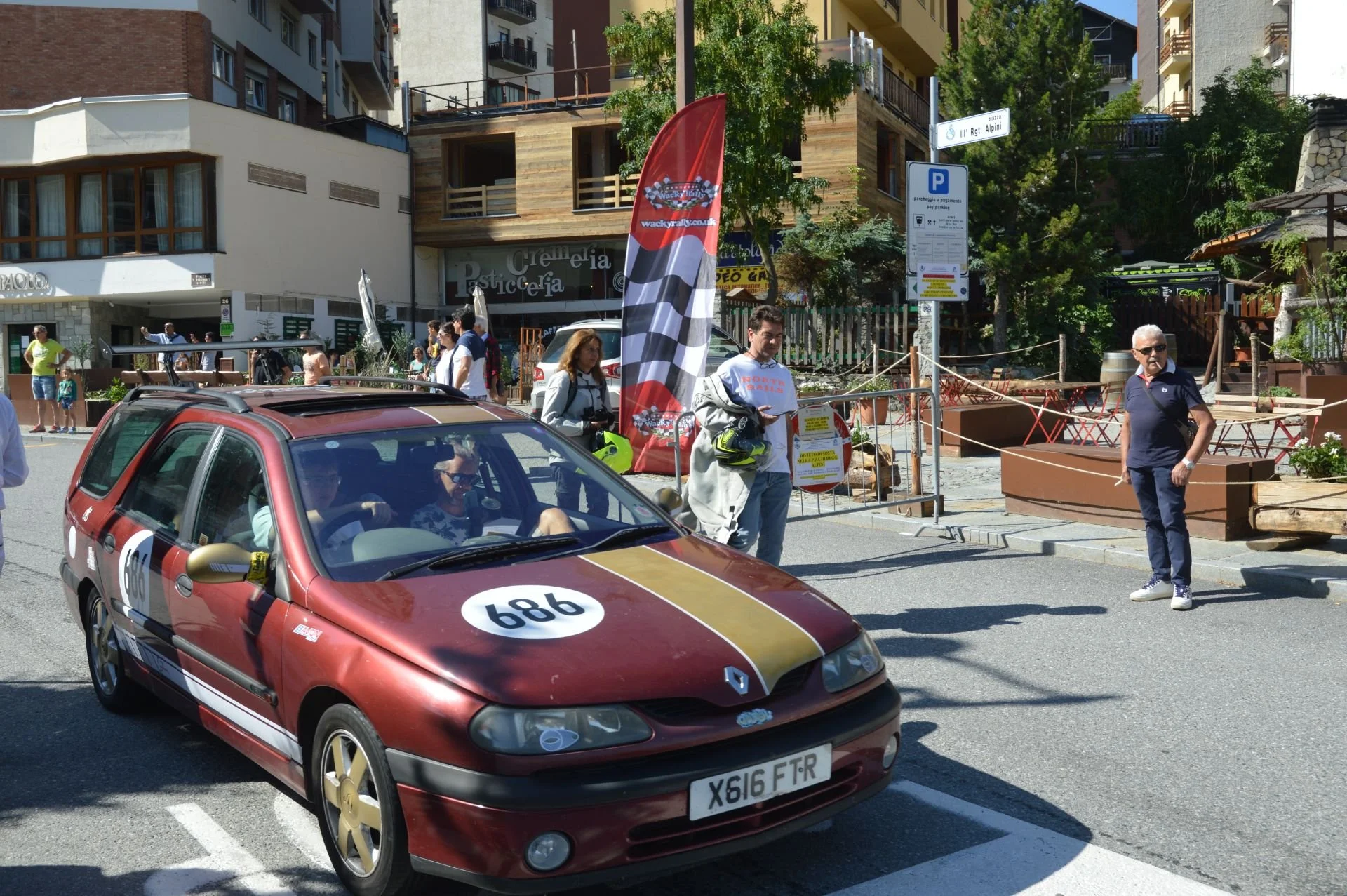 A vintage maroon Renault car with racing decals and the number 686, parked on a city street during a rally event. People are walking around, some looking at the car, and there are banners and signs related to the event in the background.