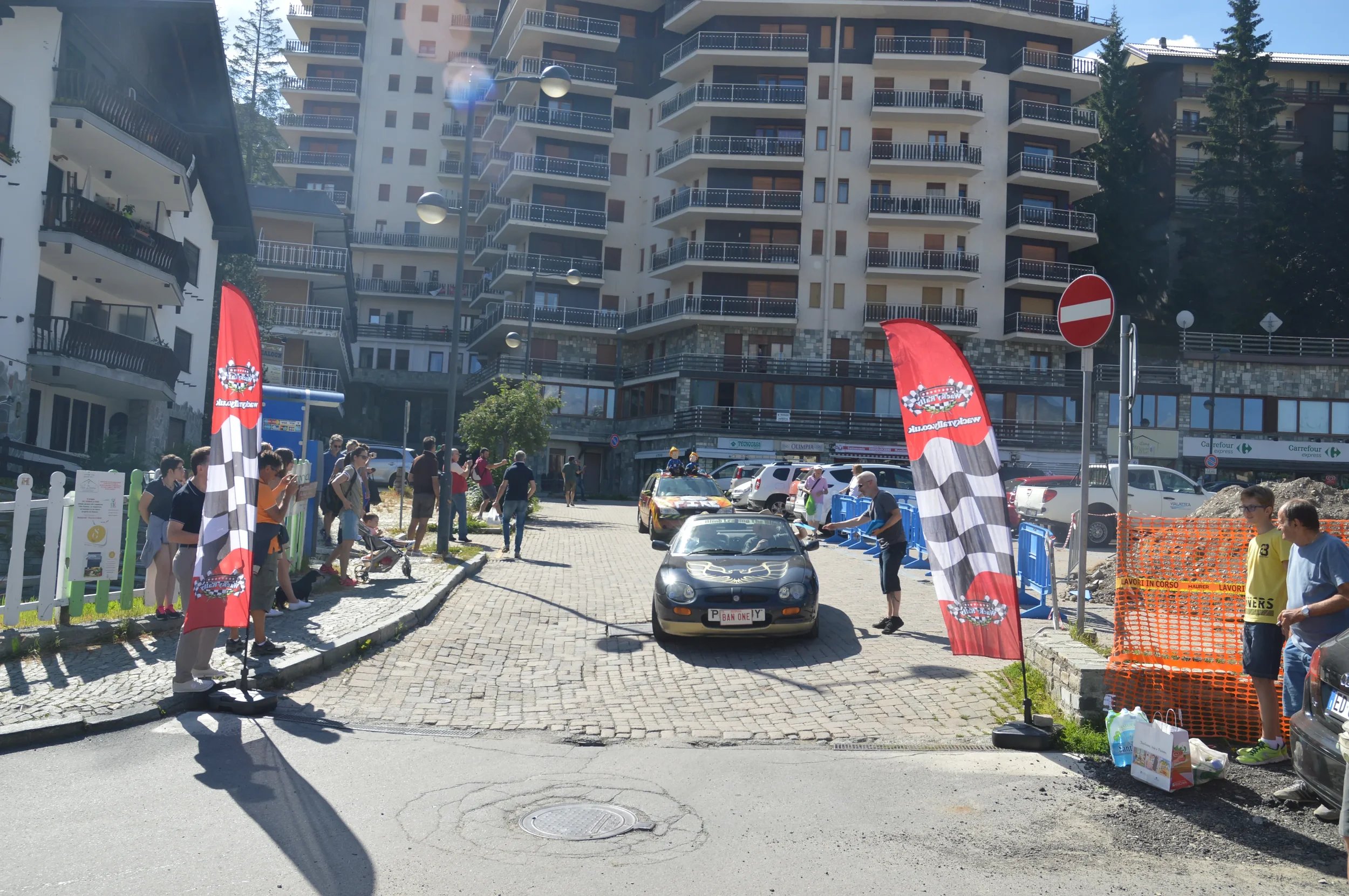 A scene of a car race event in a city street, with a sports car on a cobblestone section, surrounded by people, flags, and race officials. Tall residential buildings and trees are in the background.