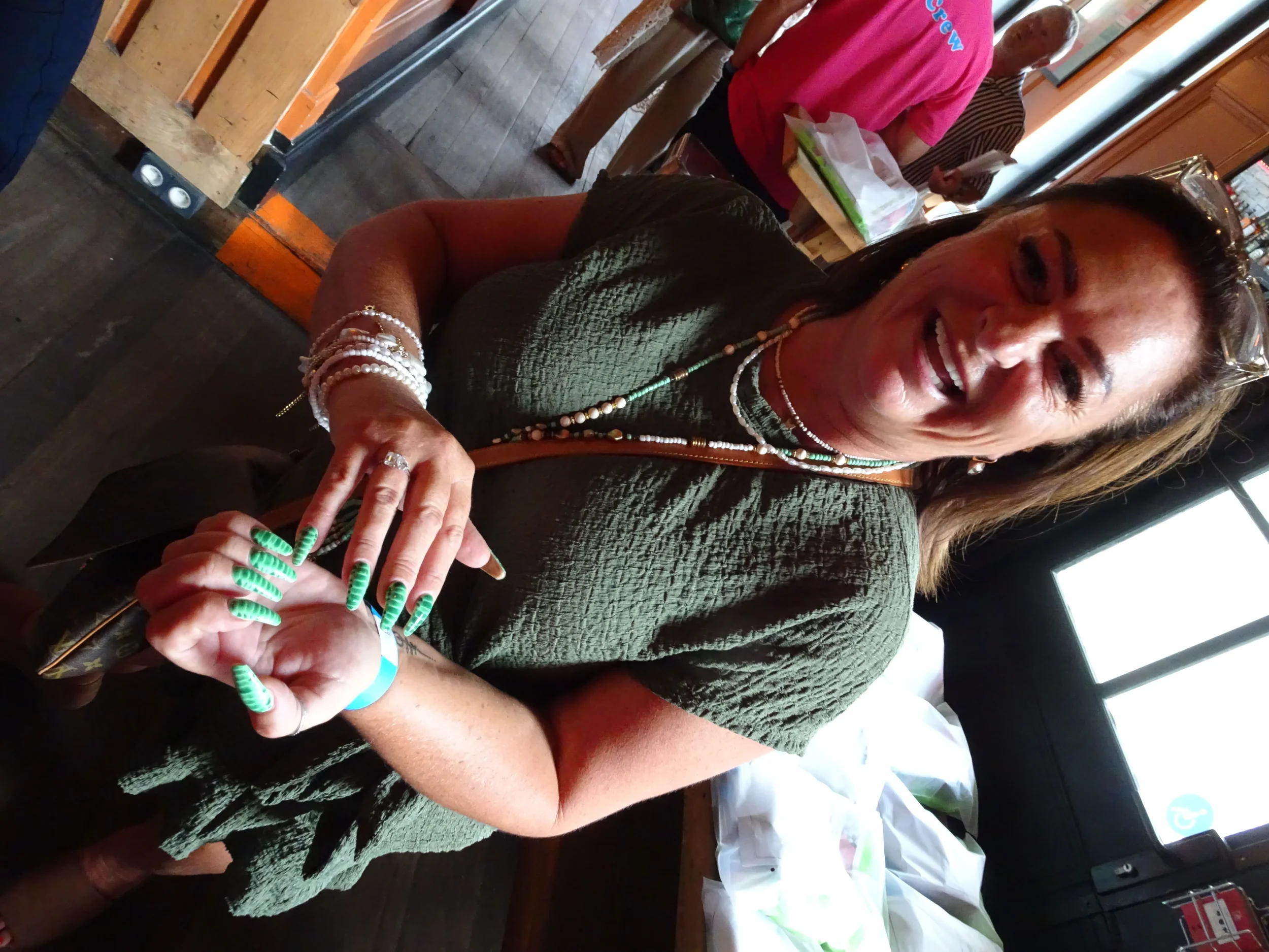 A woman with long brown hair smiling and wearing glasses on her head, dressed in a green textured top, sitting at a table with her hands clasped showing long green and white striped nails, and multiple jewelry pieces. In the background, there are a f