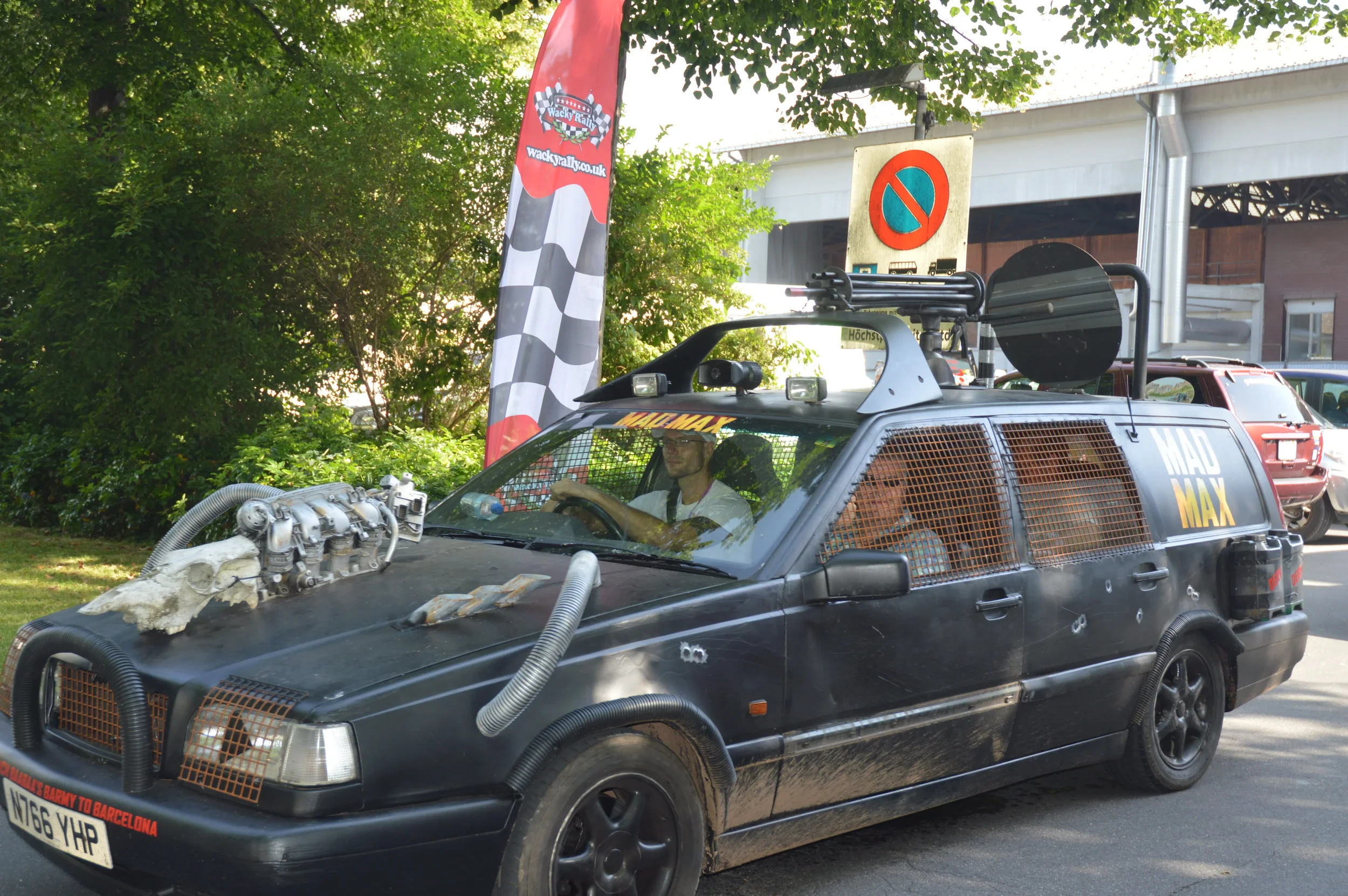 A customized black station wagon vehicle with various modifications and accessories, including a skull ornament on the hood, spray foam like material, large vents, and cage-like window guards. People are inside the car, and it is parked outdoors near