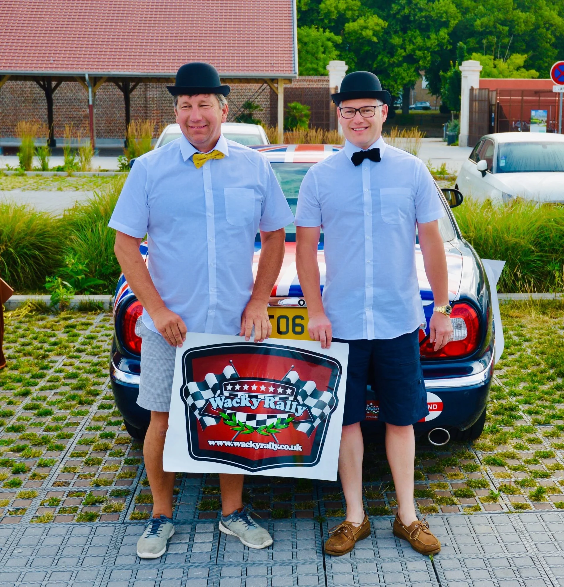 Two men dressed in light blue shirts, bow ties, and black bowler hats standing in front of a rally car, holding a sign that says "Wacky Rally" with a website link, outdoors in a parking area.