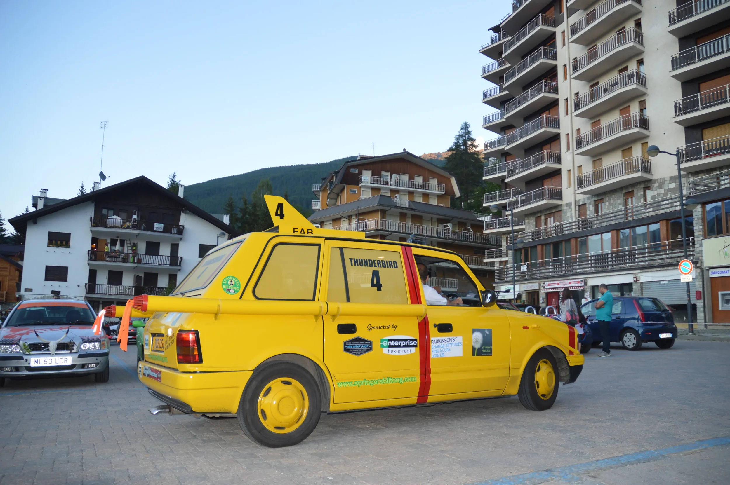 A yellow vehicle labeled 'Thunderbird 4' parked in a European town square with people walking by and multi-story buildings in the background.