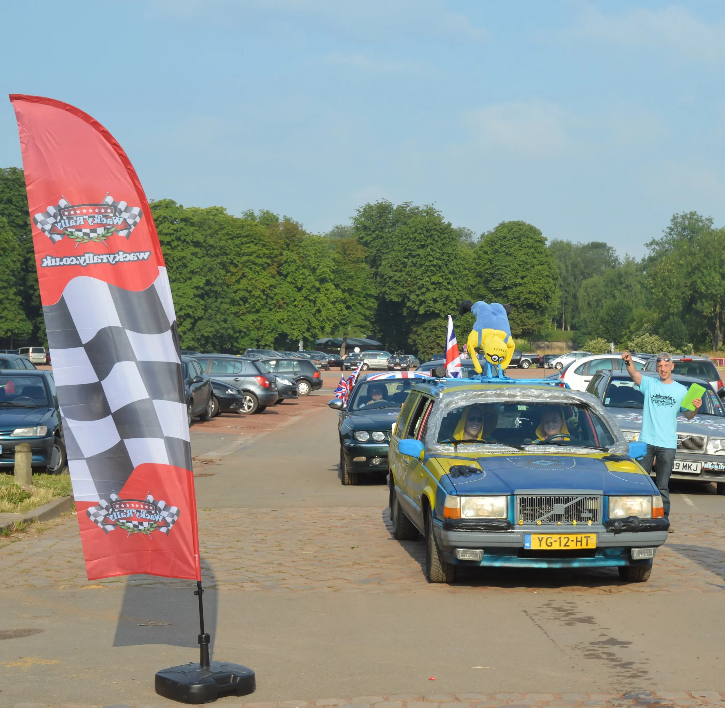 A parade of decorated cars on a parking lot with a flag, a Minion plush toy on top, and a man smiling and waving. The lot is surrounded by trees and parked cars.