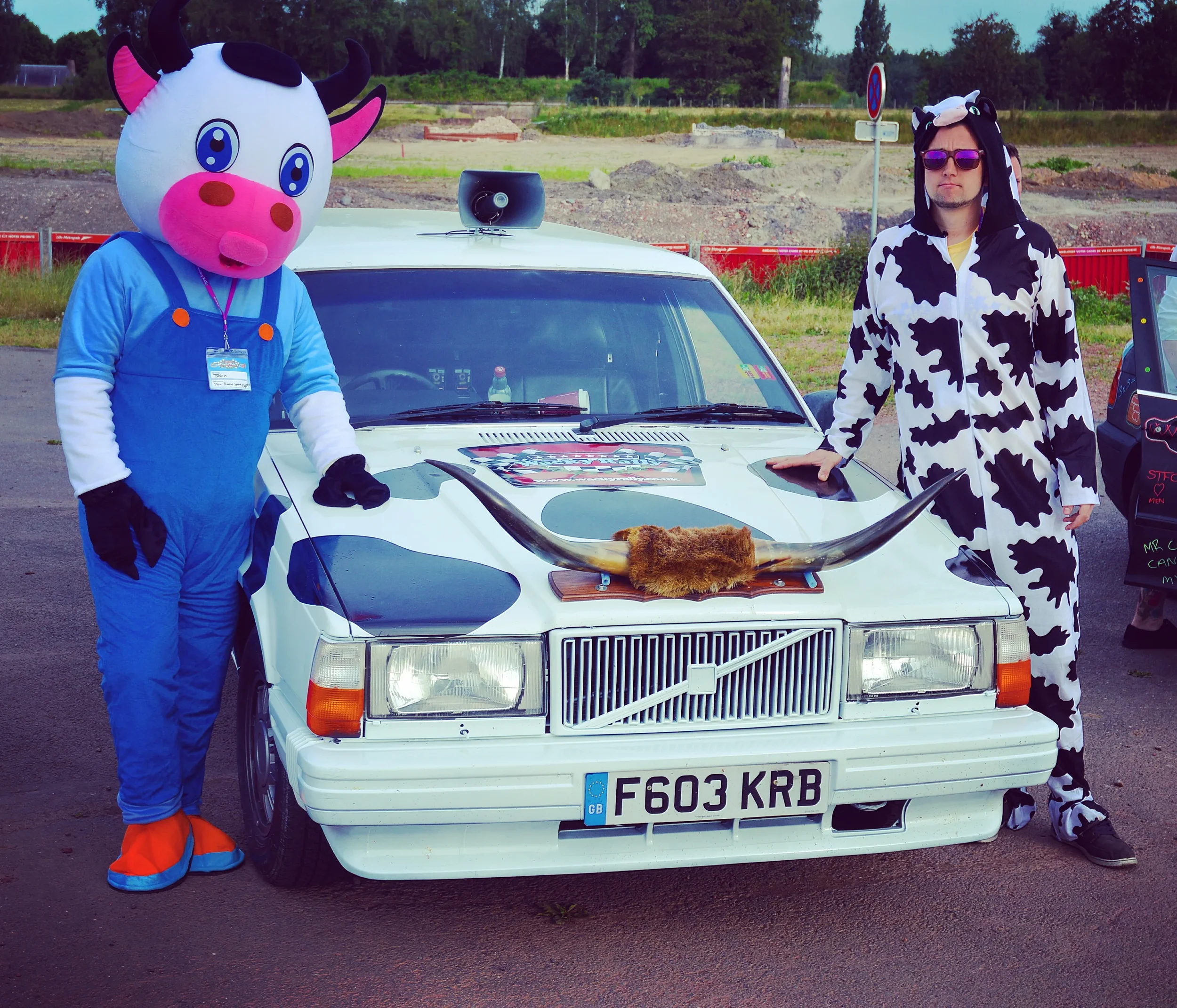 Person in a cow costume and person in a cow print onesie standing next to a white car with cow-themed decorations, including horns on the front, at an outdoor event.