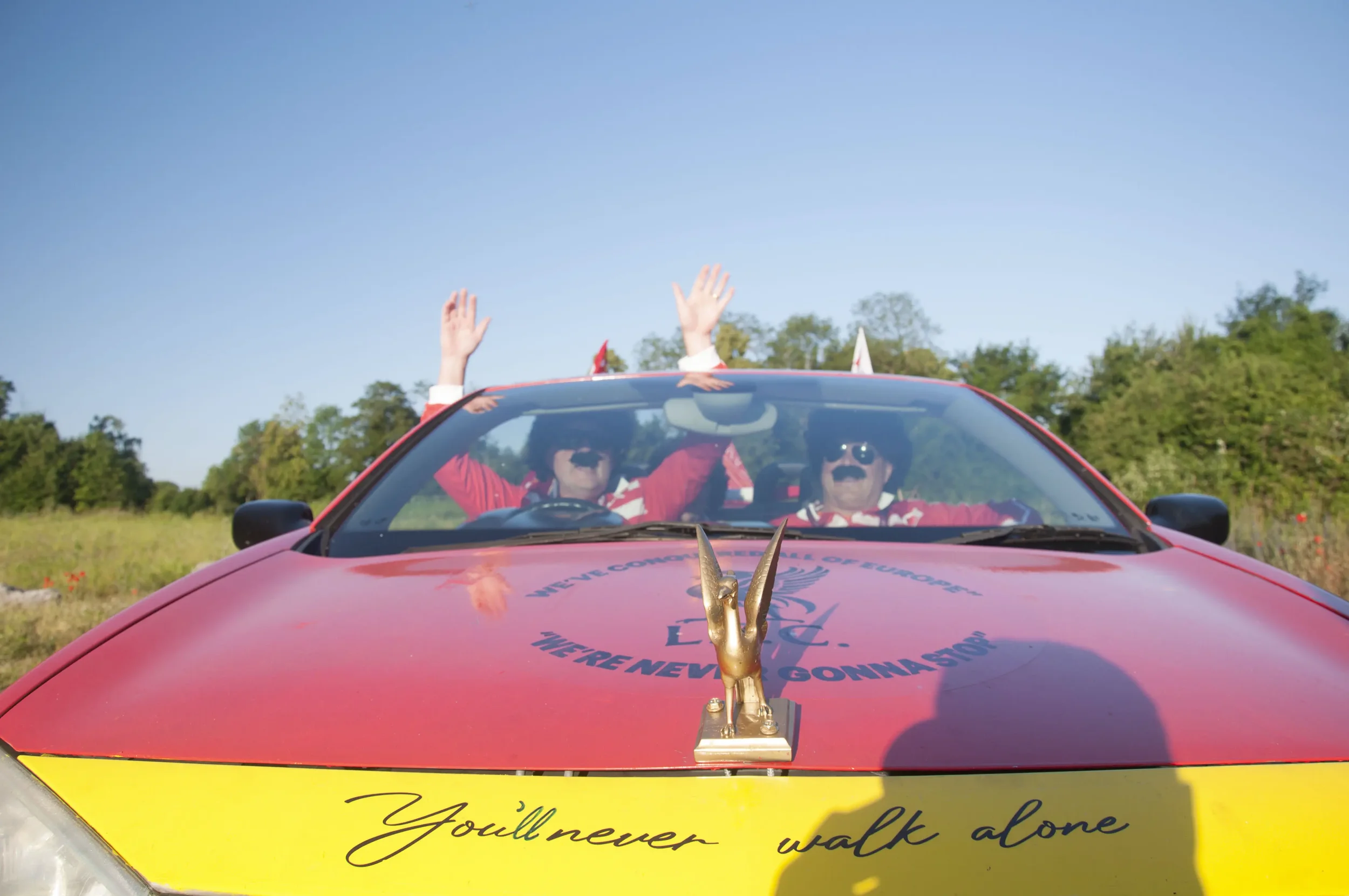 A red car with a gold rabbit sculpture on the hood, two people dressed as the Beatles with wigs, sunglasses, and mustaches inside, waving hats through the windshield, with a blue sky and green trees in the background.