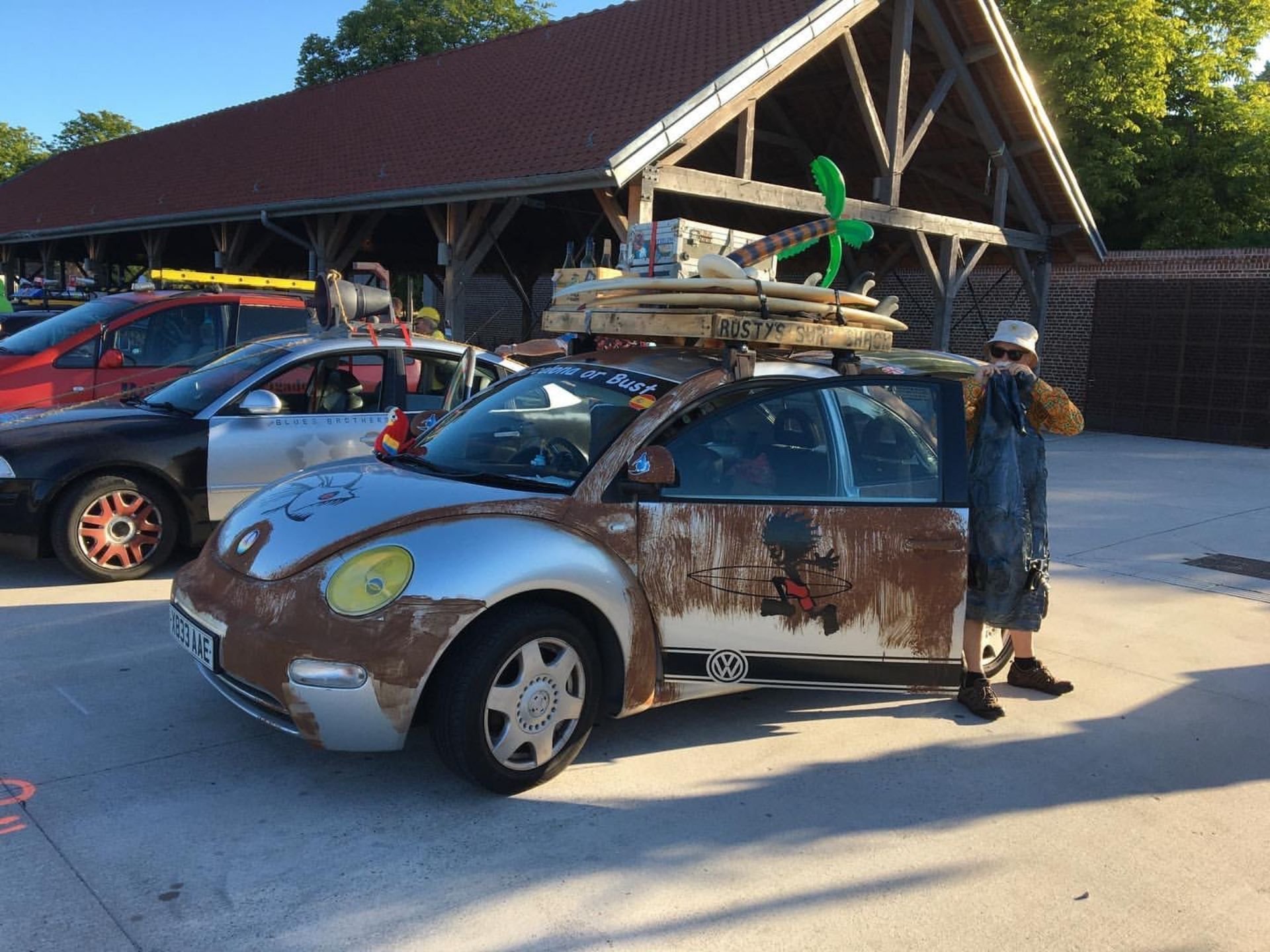 A person dressed in a patchwork outfit standing next to a Volkswagen Beetle car decorated with artistic graphics. The car has surfboards and other beach gear on the roof. A police car is parked nearby, and a rustic building with a large roof is in th