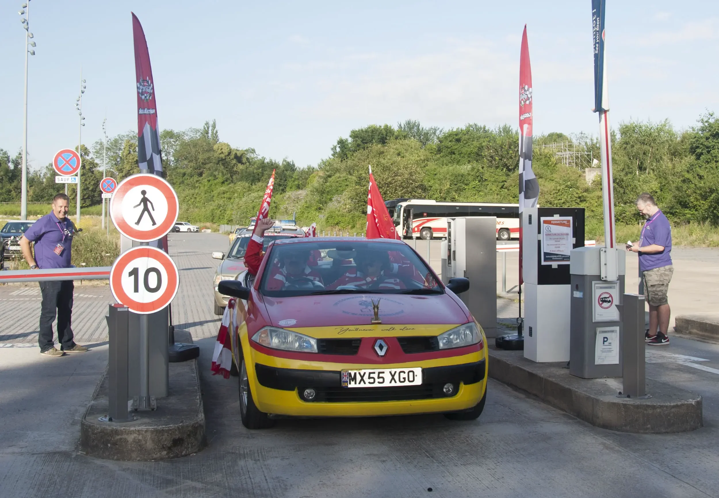 A red and yellow Renault car with flags on it is passing through a parking gate. Two signs indicate pedestrian crossing and a 10 km/h speed limit. Two men with purple shirts are on either side of the gate, one smiling and the other looking at his pho