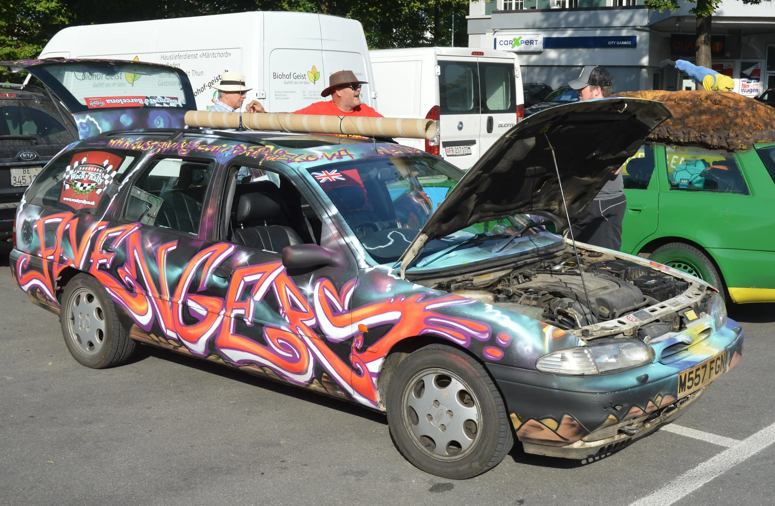 A colorful, graffiti-style decorated car with the hood open revealing the engine, parked in a lot among other vehicles and people.