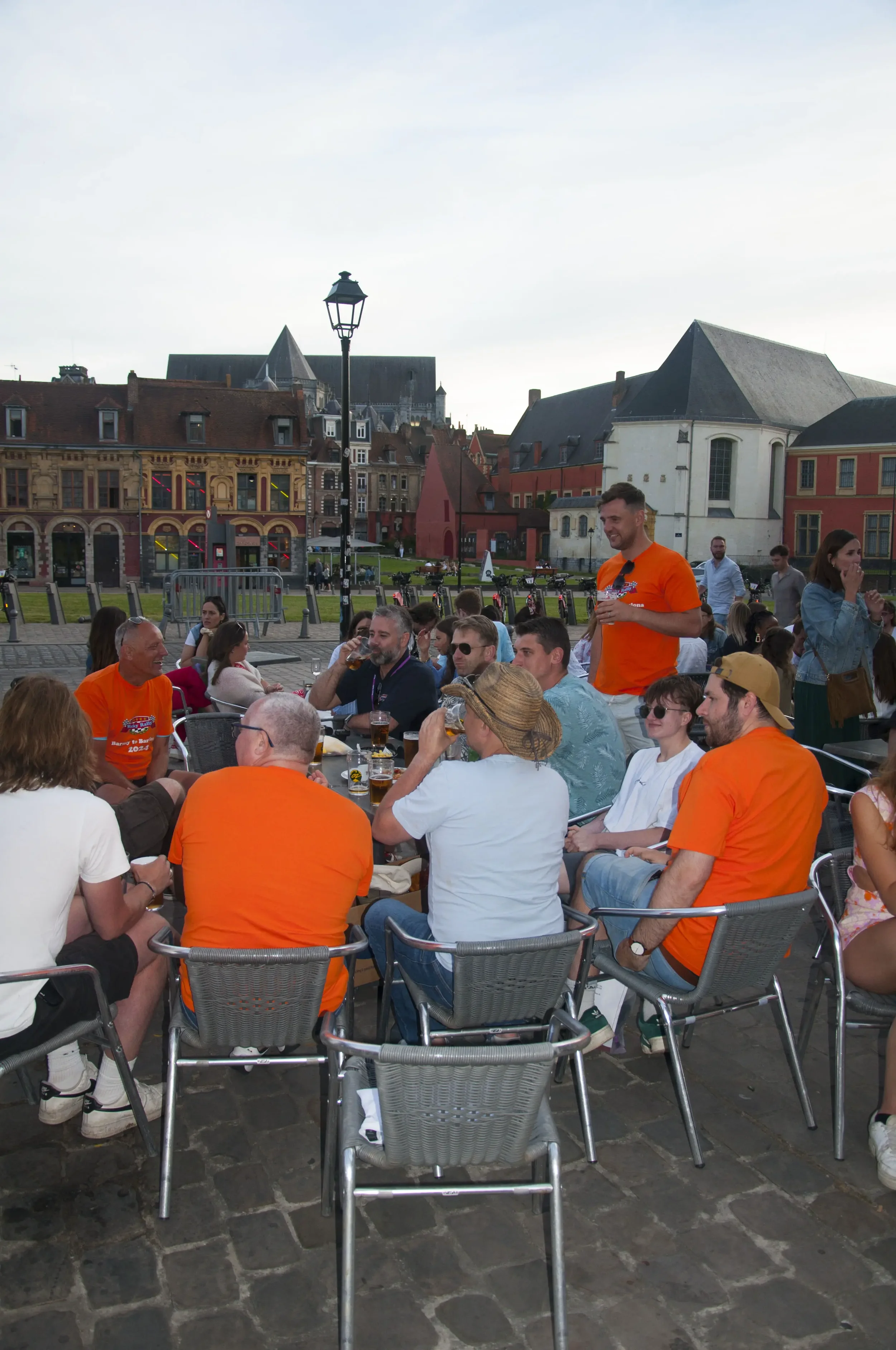 People gathered around a table outdoors in a city square, some wearing orange shirts, with historic buildings and a cloudy sky in the background.