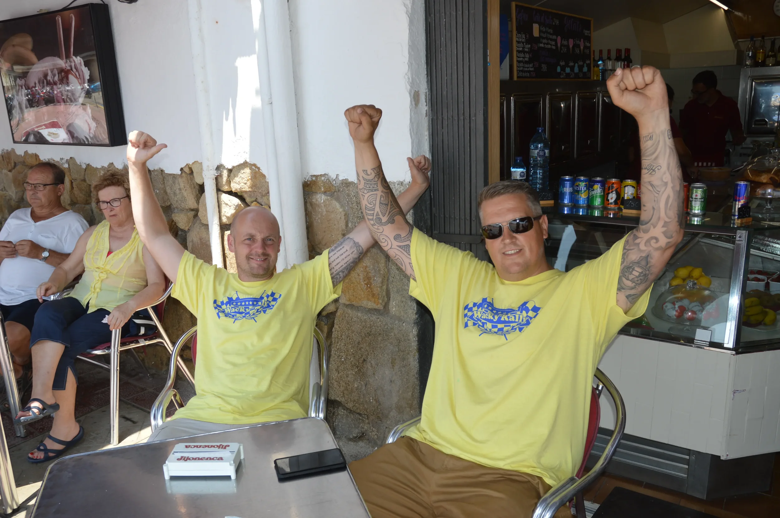 Two men wearing matching yellow shirts with graphic designs sit at a table, celebrating with raised fists and smiling, with others sitting behind them in an outdoor cafe or restaurant setting.
