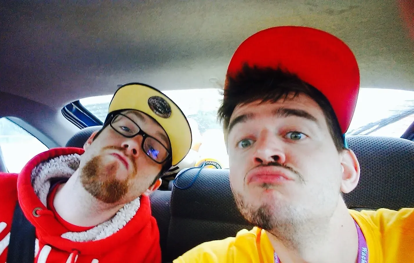 Two young men sitting in the backseat of a car, taking a selfie. The man on the left has glasses, a beard, and is wearing a yellow hat and a red hoodie. The man on the right has a red hat, light skin, and is wearing a yellow shirt.
