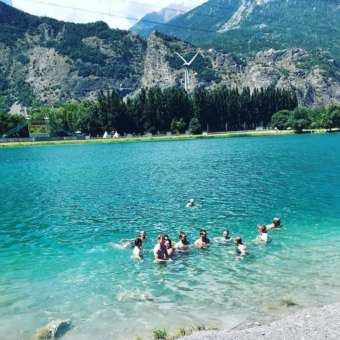 People swimming in a clear, blue lake with a mountainous background and a wind turbine on the mountain.