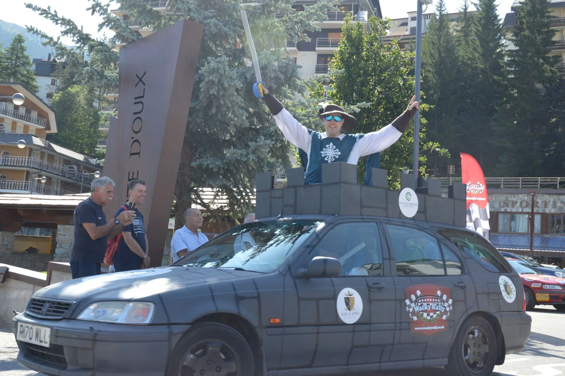 A person dressed as a king with a crown, sunglasses, and a cape, standing in a cardboard castle atop a small car, holding a sword in the air during a rally or parade. Several onlookers are watching and taking pictures nearby.