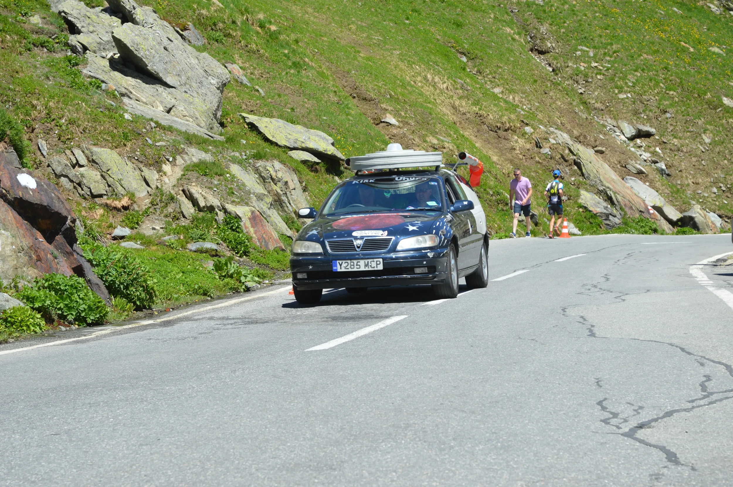 A black car parked on the side of a mountain road with a mural of the American flag on its hood, while two people walk nearby and another person with a backpack brushes the area.