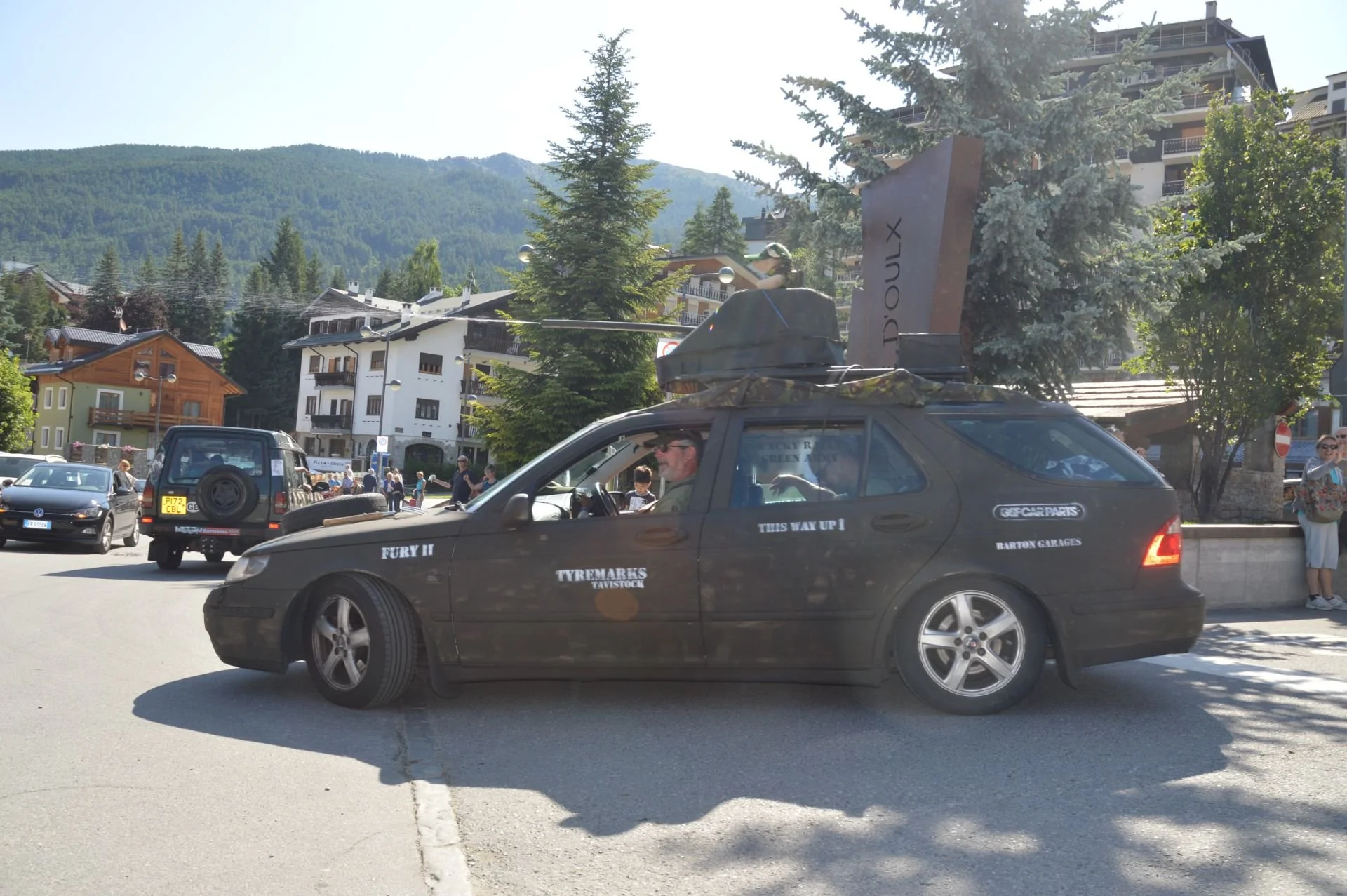 Black station wagon with decals and a rooftop display, parked on a street in a mountainous town with buildings, trees, and people in the background.