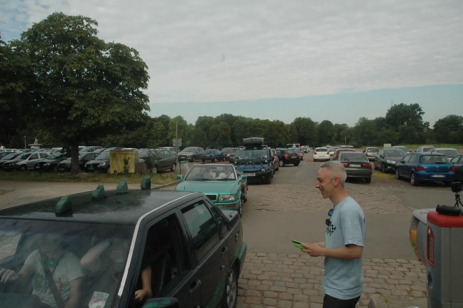 A parking lot filled with cars, with a man standing on the sidewalk holding a smartphone and smiling, a man sitting in a black car, a tree on the left, and cloudy skies overhead.