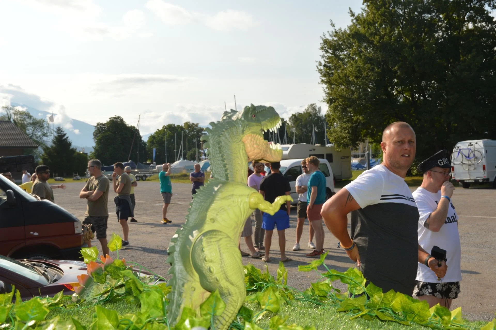 A large green inflatable dinosaur on display outdoors at a gathering, with people in casual clothing standing and talking in the background near parked cars, boats, and trees.