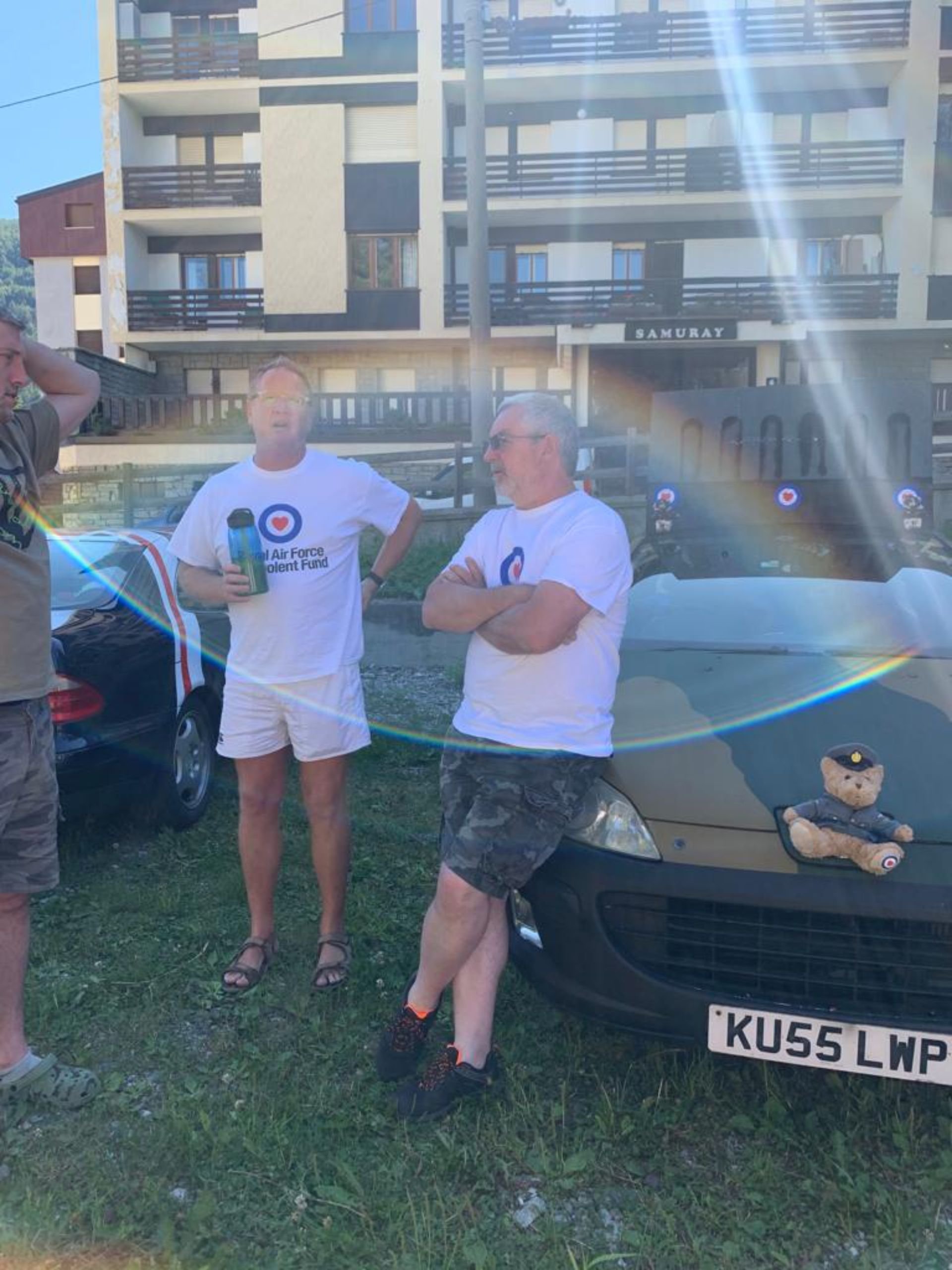 Three men standing outdoors near a car, with one holding a water bottle, wearing shirts with a logo and text referencing the Royal Air Force. A teddy bear dressed as a policeman sits on the car's front grille. In the background, there is a modern mul