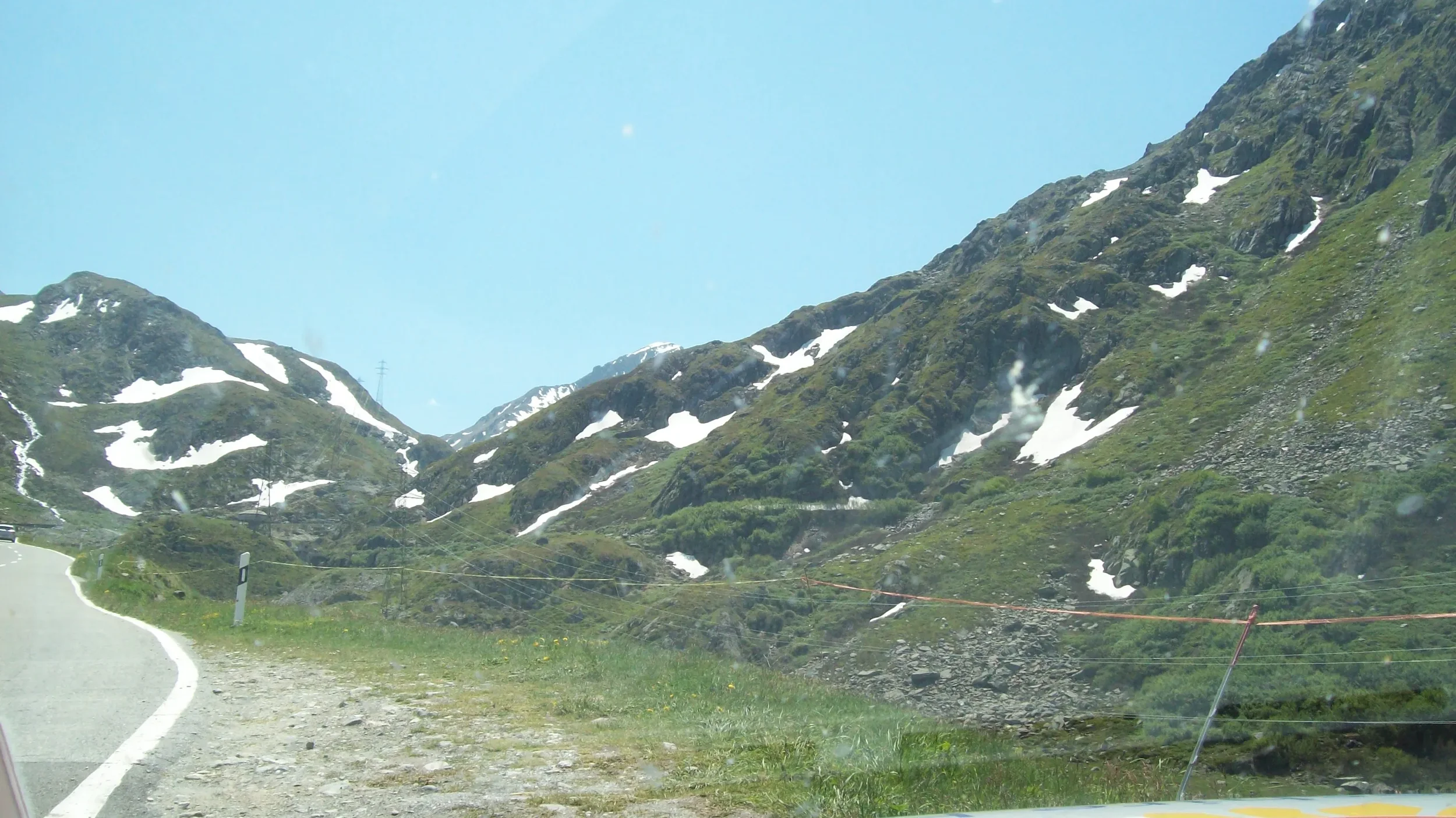 Mountain landscape with patches of snow, green vegetation, and a winding road with a safety barrier.
