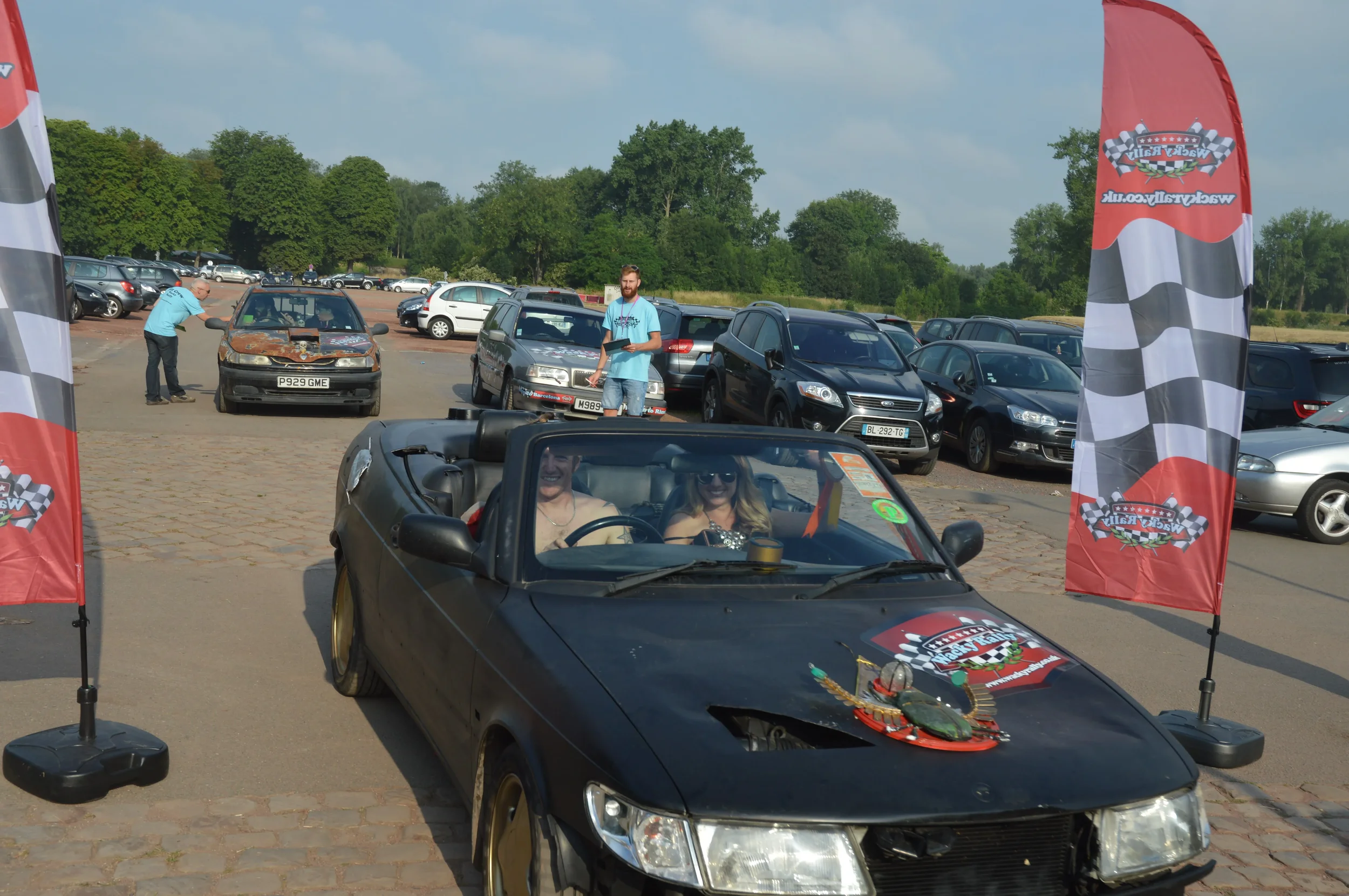 Black convertible car decorated with holiday ornaments and a sign, with two women inside, parked at an outdoor car event with flags and other cars around.