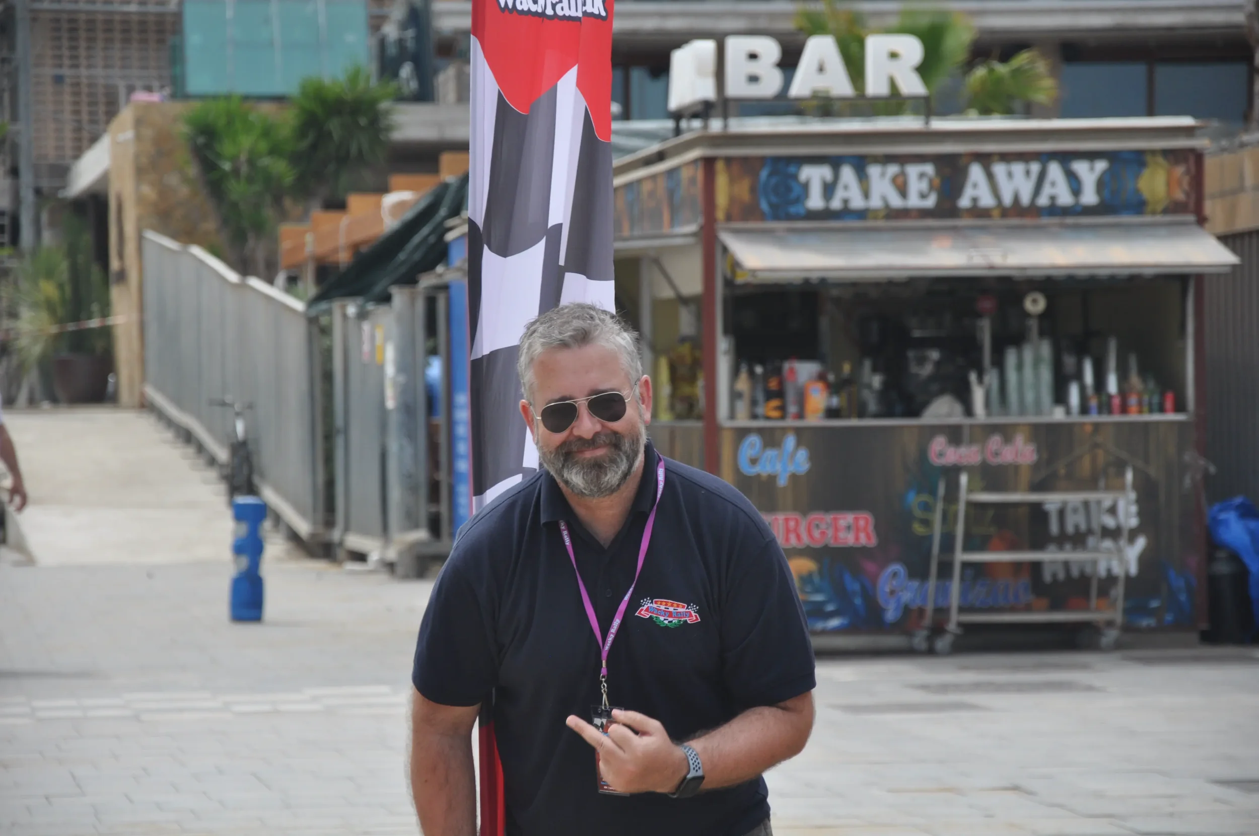 A man with gray hair, beard, sunglasses, wearing a black polo shirt with a logo, standing outdoors near a cafe with a take away stand, flags, and some greenery.