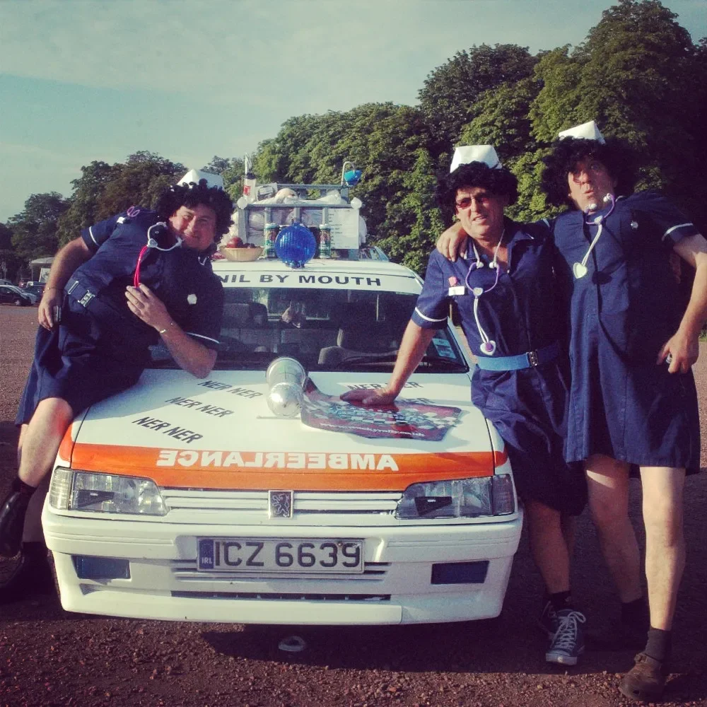 Three men dressed as nurses posing with an ambulance car decorated with medical props outside, with trees in the background.