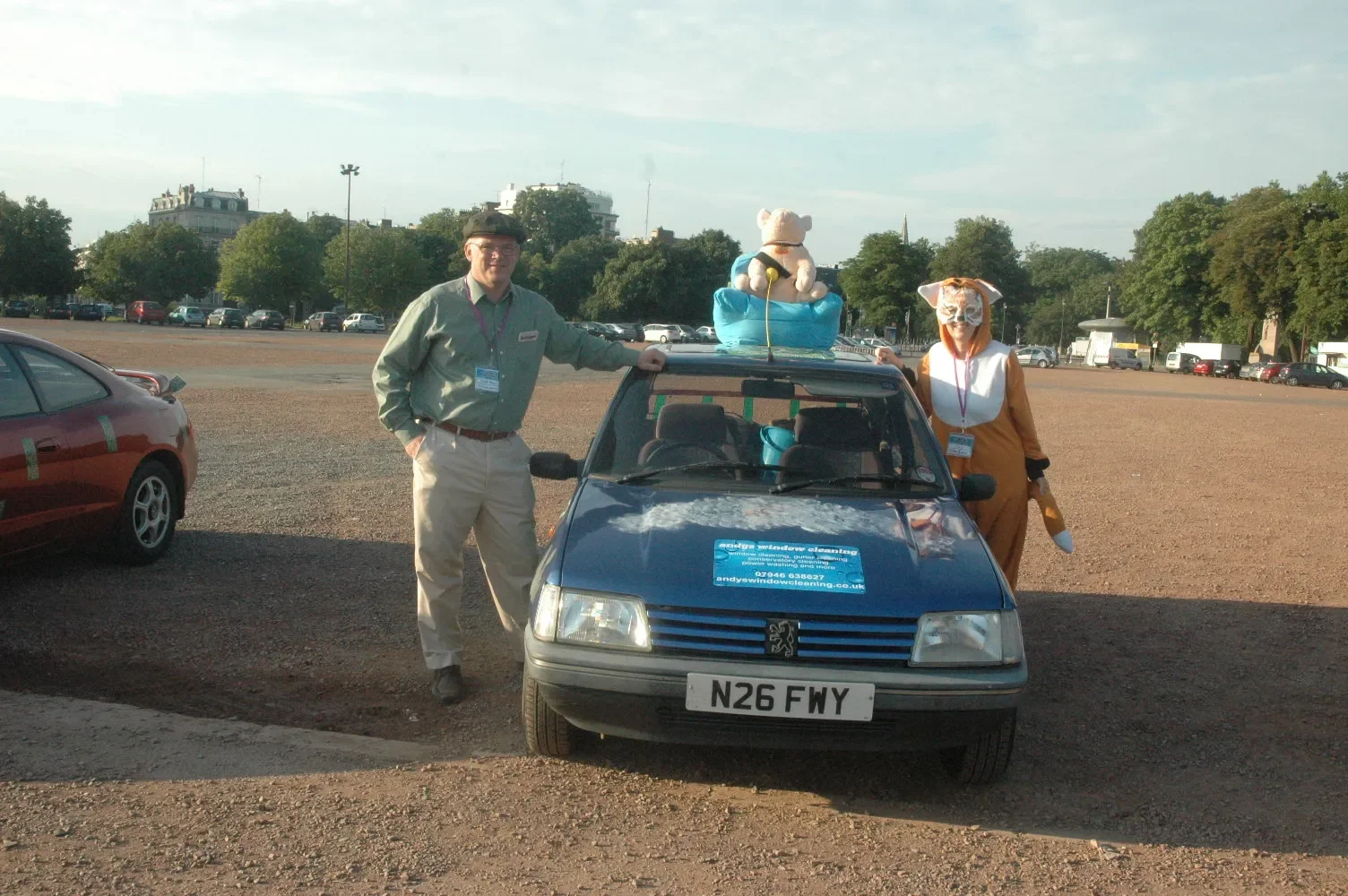 A man and a woman in costumes are standing beside a small Peugeot car decorated with a sign and stuffed animals on top, in a parking lot with trees and buildings in the background.