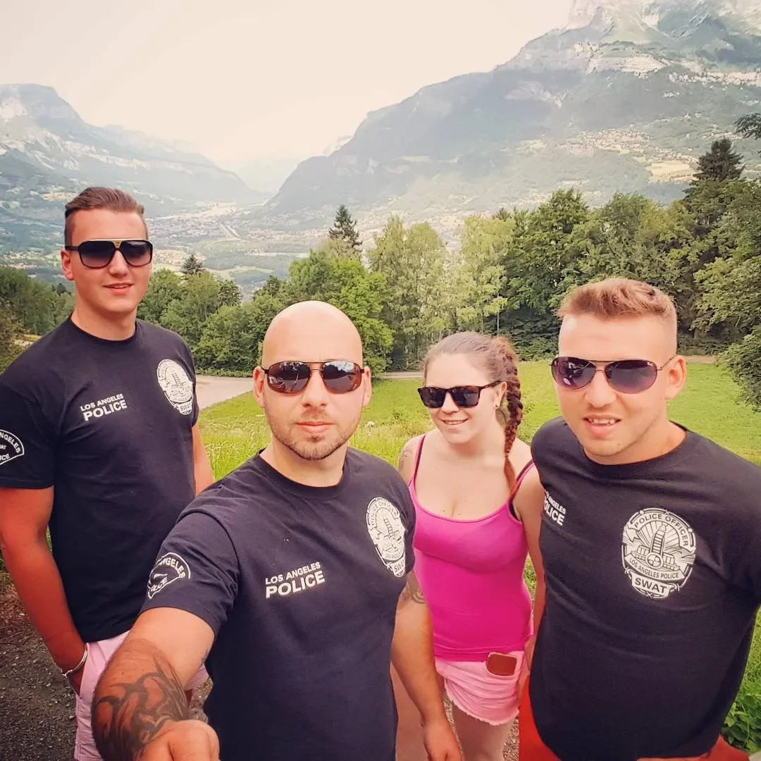 Four people standing outdoors with mountains and trees in the background. Three men and one woman, all wearing sunglasses. Two men are in black police or SWAT shirts, and the woman is in a pink top and shorts.