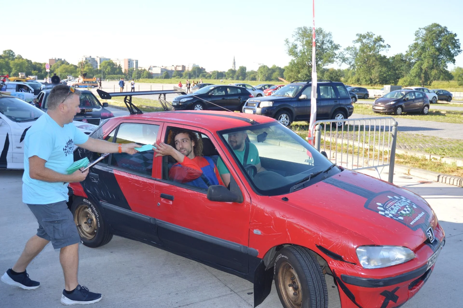 A man in a light blue t-shirt and shorts is handing a stack of papers to a person inside a red and black car at a daytime outdoor event.