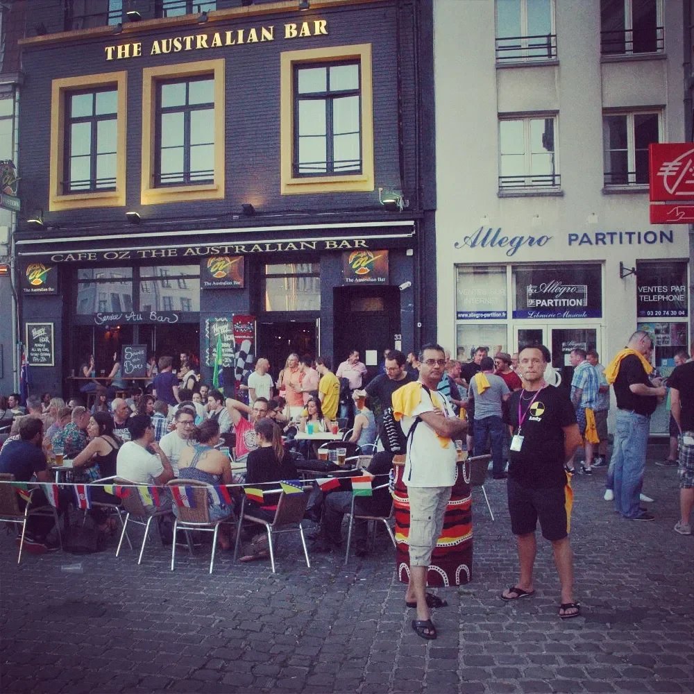 An outdoor scene in front of a bar called 'The Australian Bar' with many people sitting at tables and standing, some wearing Australian-themed clothing or colors, on a cobblestone street during daytime.