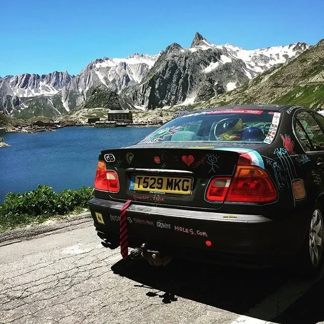 Black car with colorful graffiti parked near a lake with mountains and snow-capped peaks in the background.
