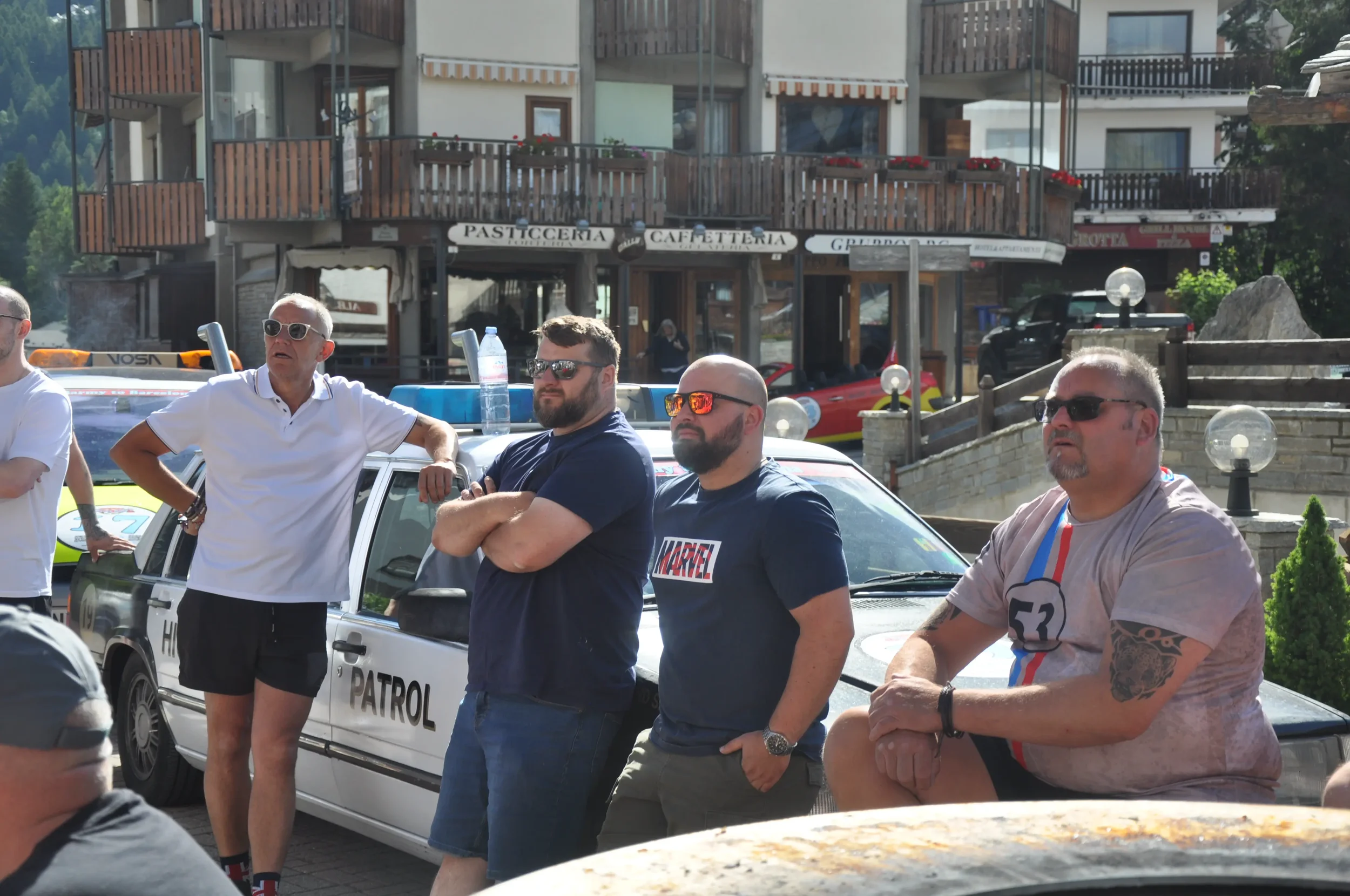 Four men standing near a patrol car in a mountainous outdoor setting, with buildings and a cafe in the background, during daytime.