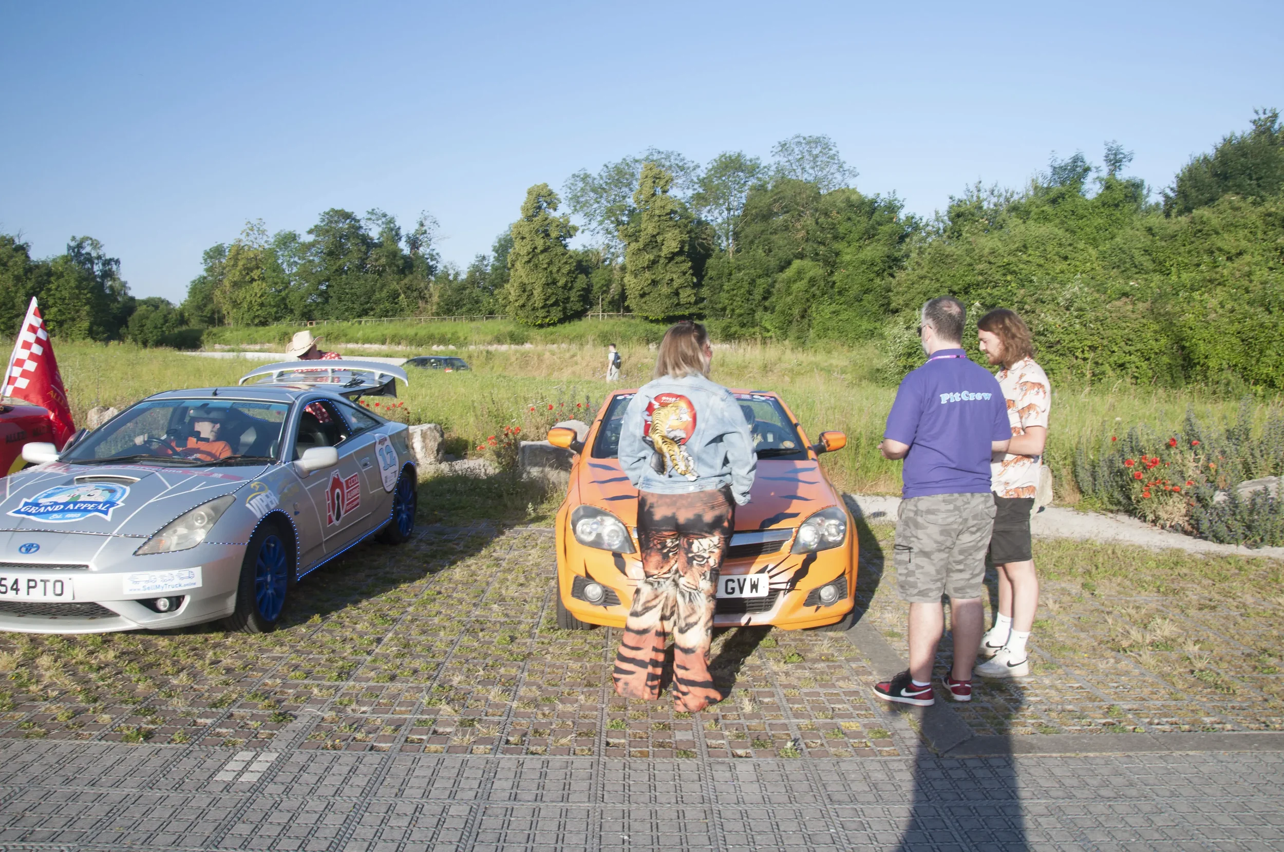 Two vintage rally cars, one with anime-themed paint, parked on a brick-paved area with grass and trees in the background. Three people stand near the cars, engaged in conversation, during daytime.