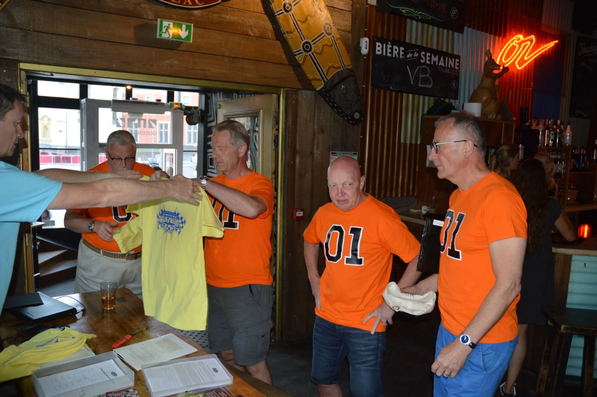 Four men in orange shirts with number 01 and 11 and one man in a blue shirt are inside a bar. One man in a blue shirt is handing a yellow T-shirt to a man in an orange shirt with number 11. The other men are watching. The bar has wooden walls, signs,