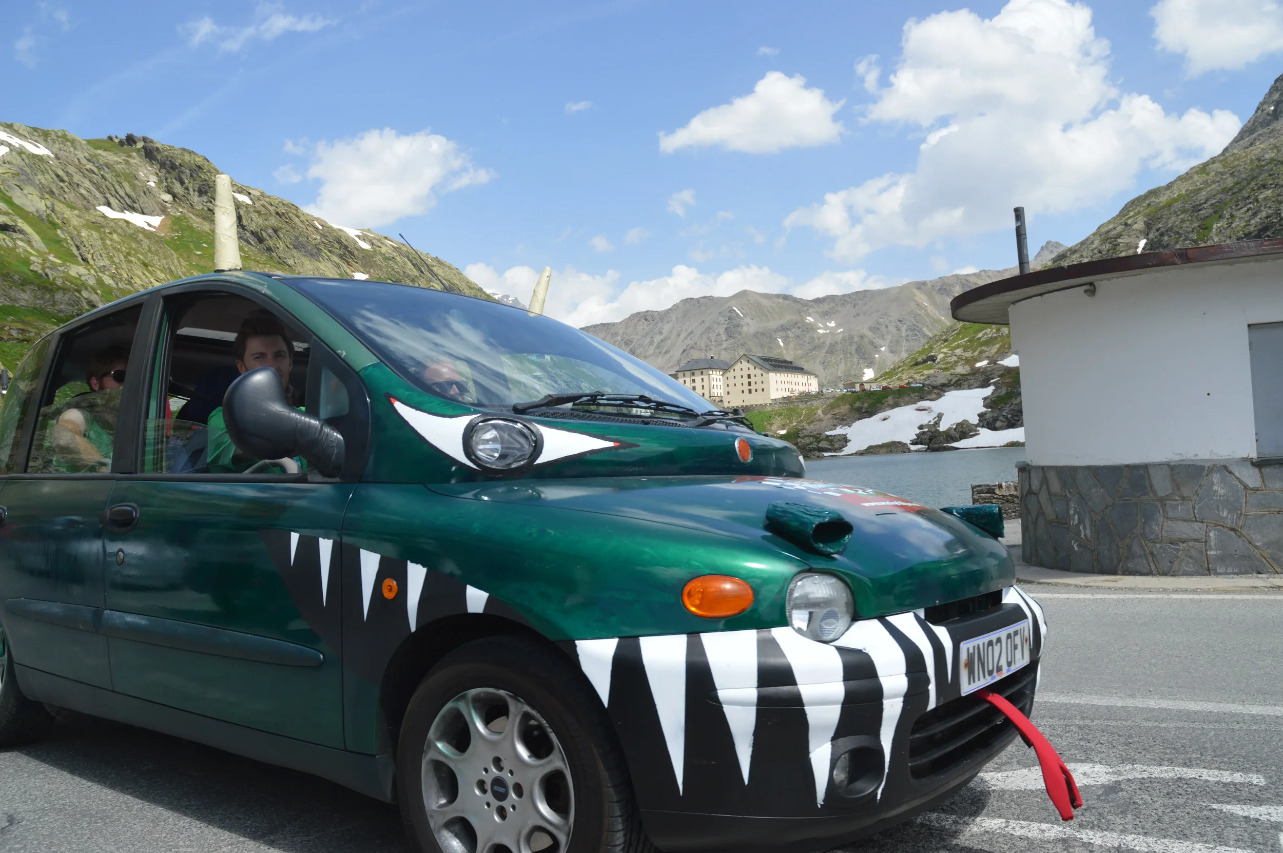 A small green car decorated to look like a shark, with painted teeth and eyes, parked on a mountain road by a lake, with mountains and a building in the background.