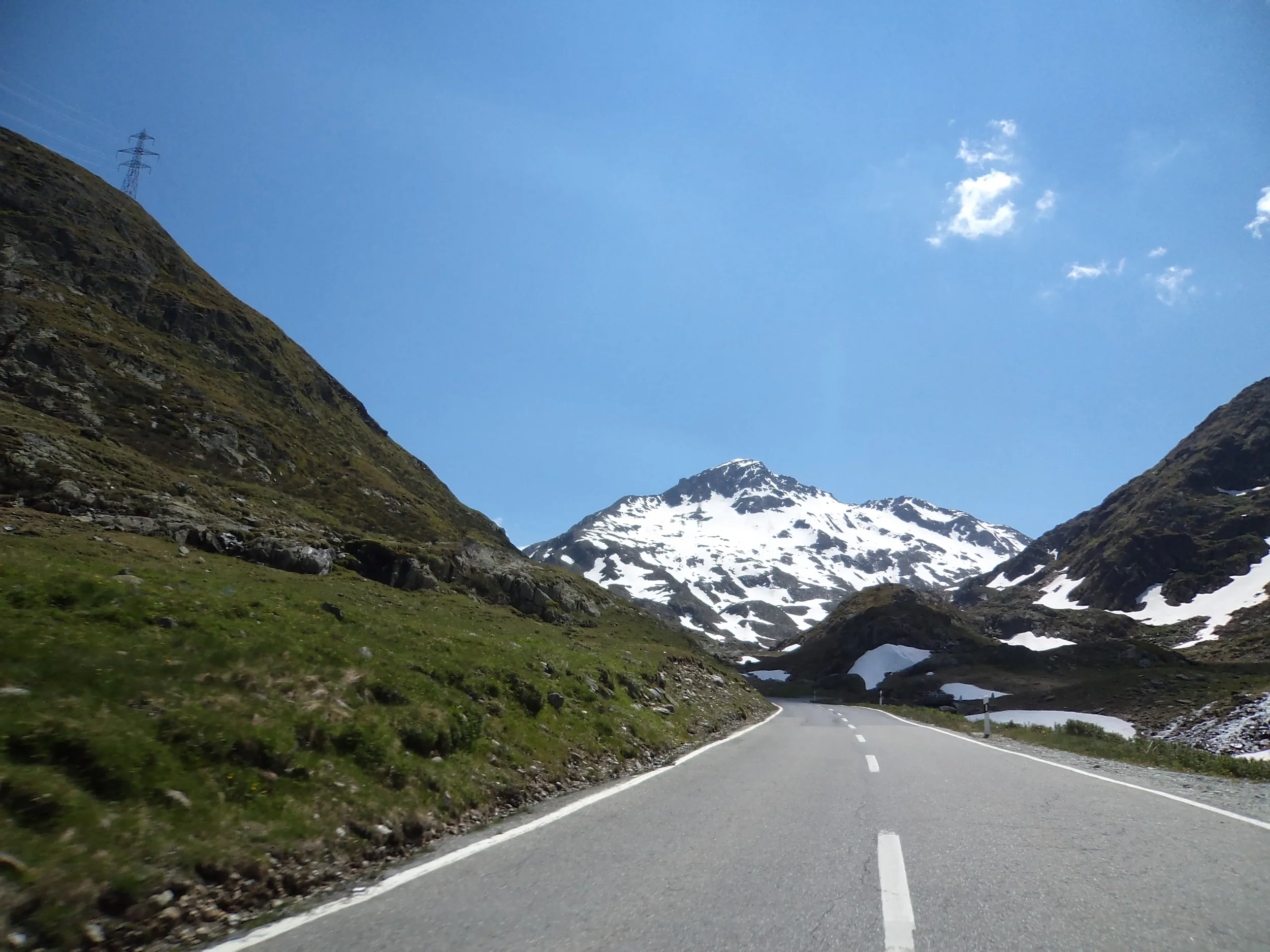 A road winding through mountainous terrain with snow-capped peaks and green slopes under a clear blue sky.