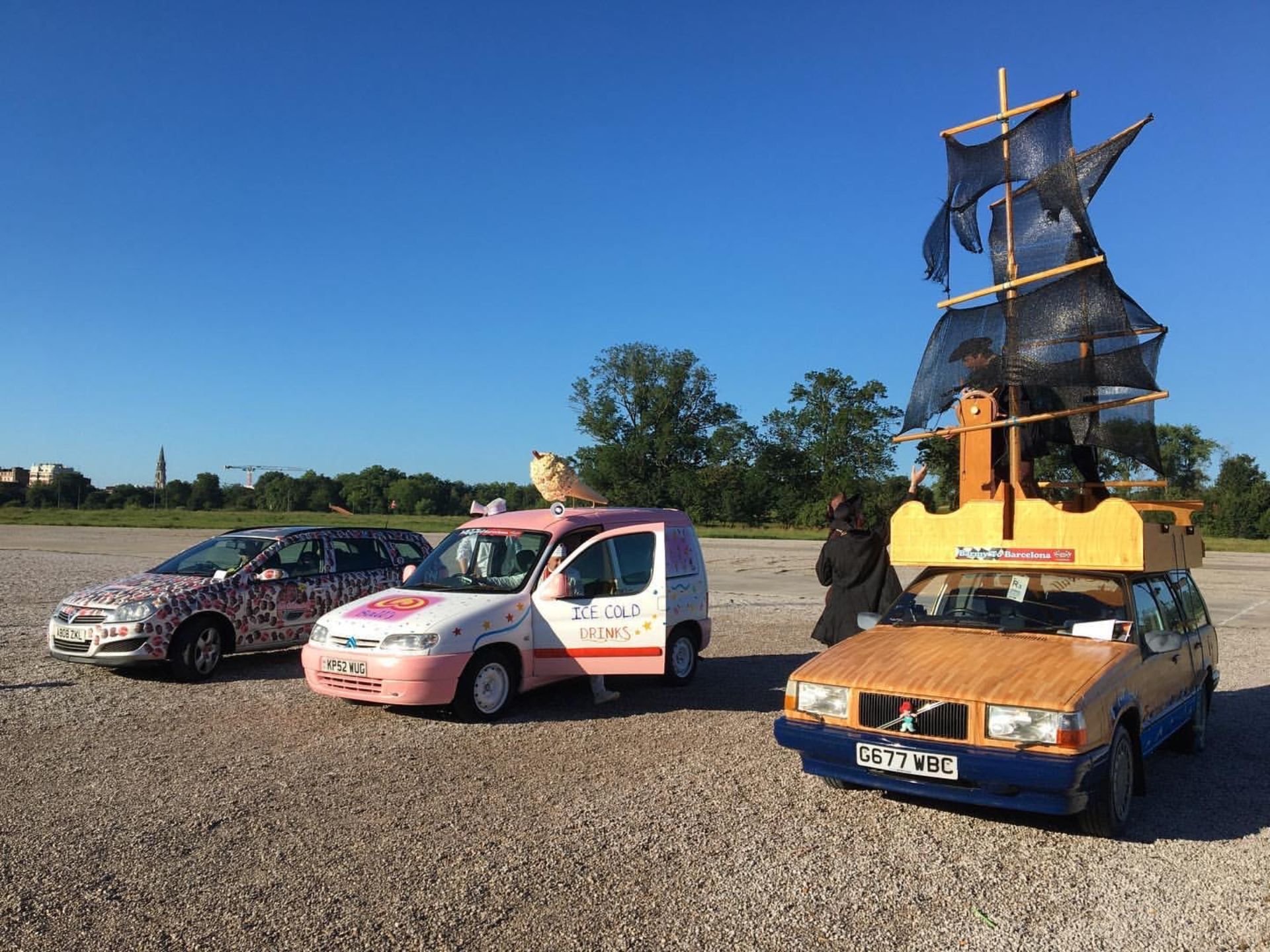 Three cars decorated with creative themes parked outdoors with a clear blue sky. The first car has a leopard print design, the second is painted with pastel colors and has a sign for ice-cold drinks, topped with a plush animal, and the third, designe